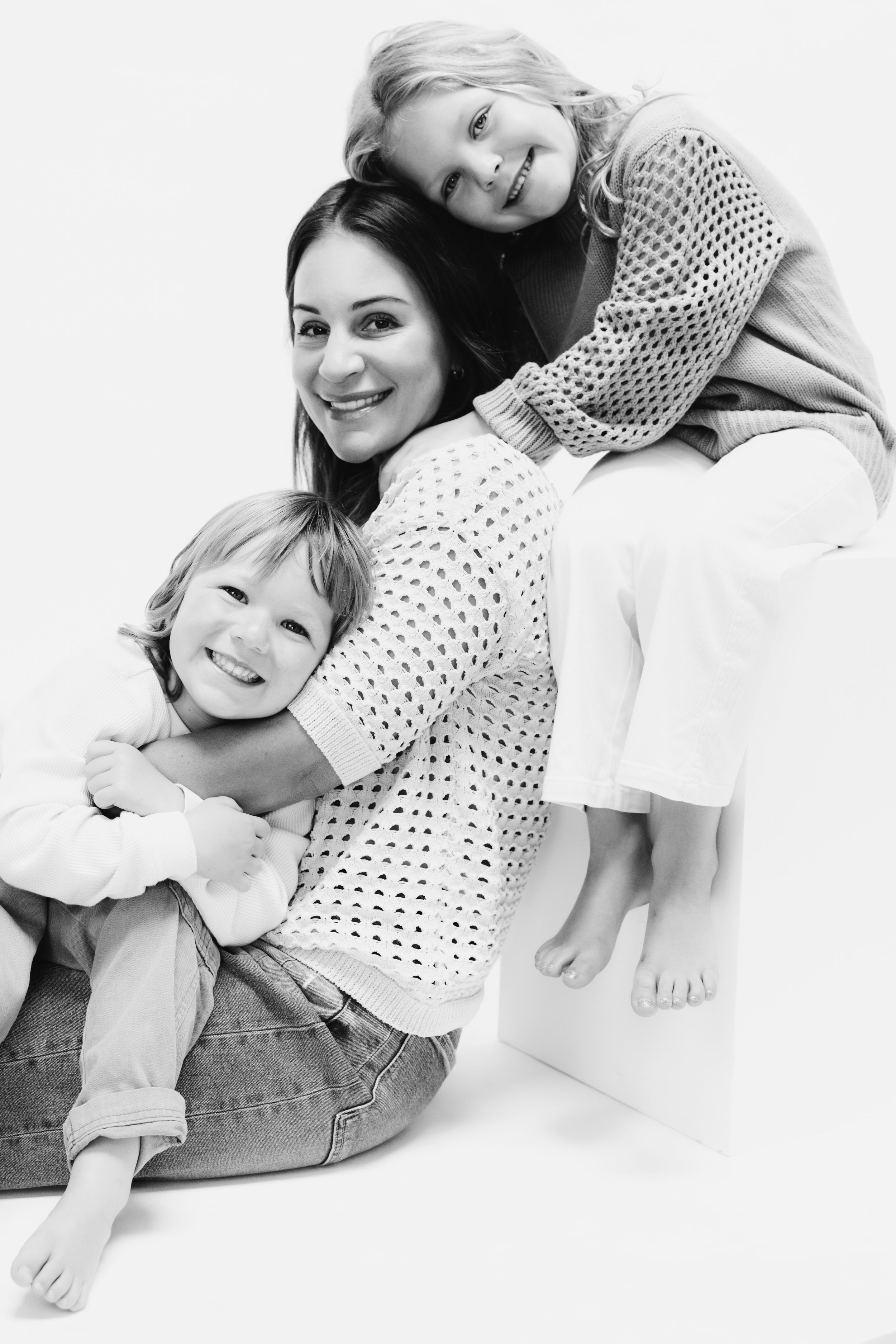 A woman with two children smiling, one sitting on her lap and the other sitting on a white cube, all hugging and smiling, in black and white.