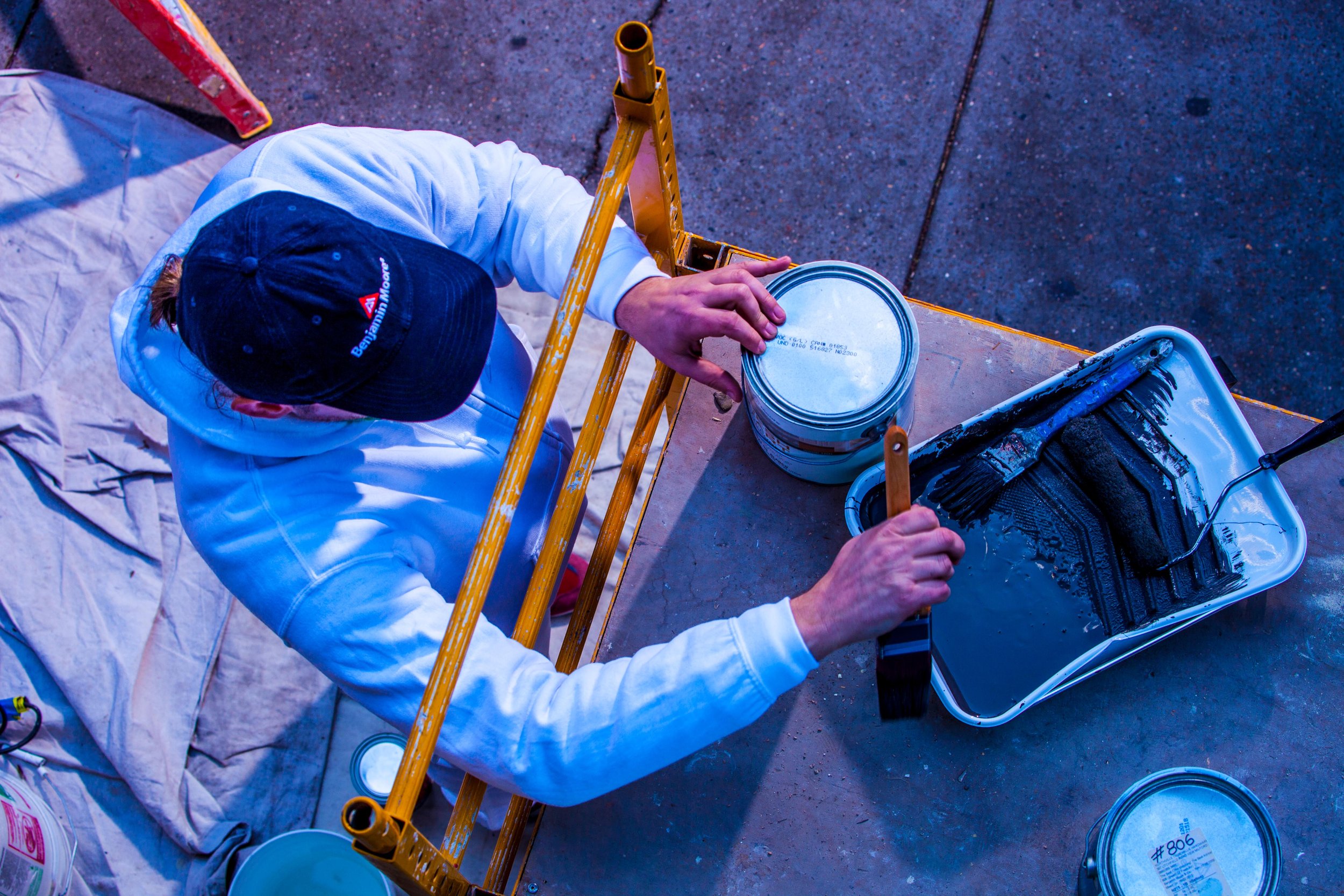 Person painting a surface with black paint, using a brush and a roller, while sitting at a work table. The person is wearing a white hoodie and a black cap with a logo. Painting supplies, including cans and a tray with black paint, are on the table.