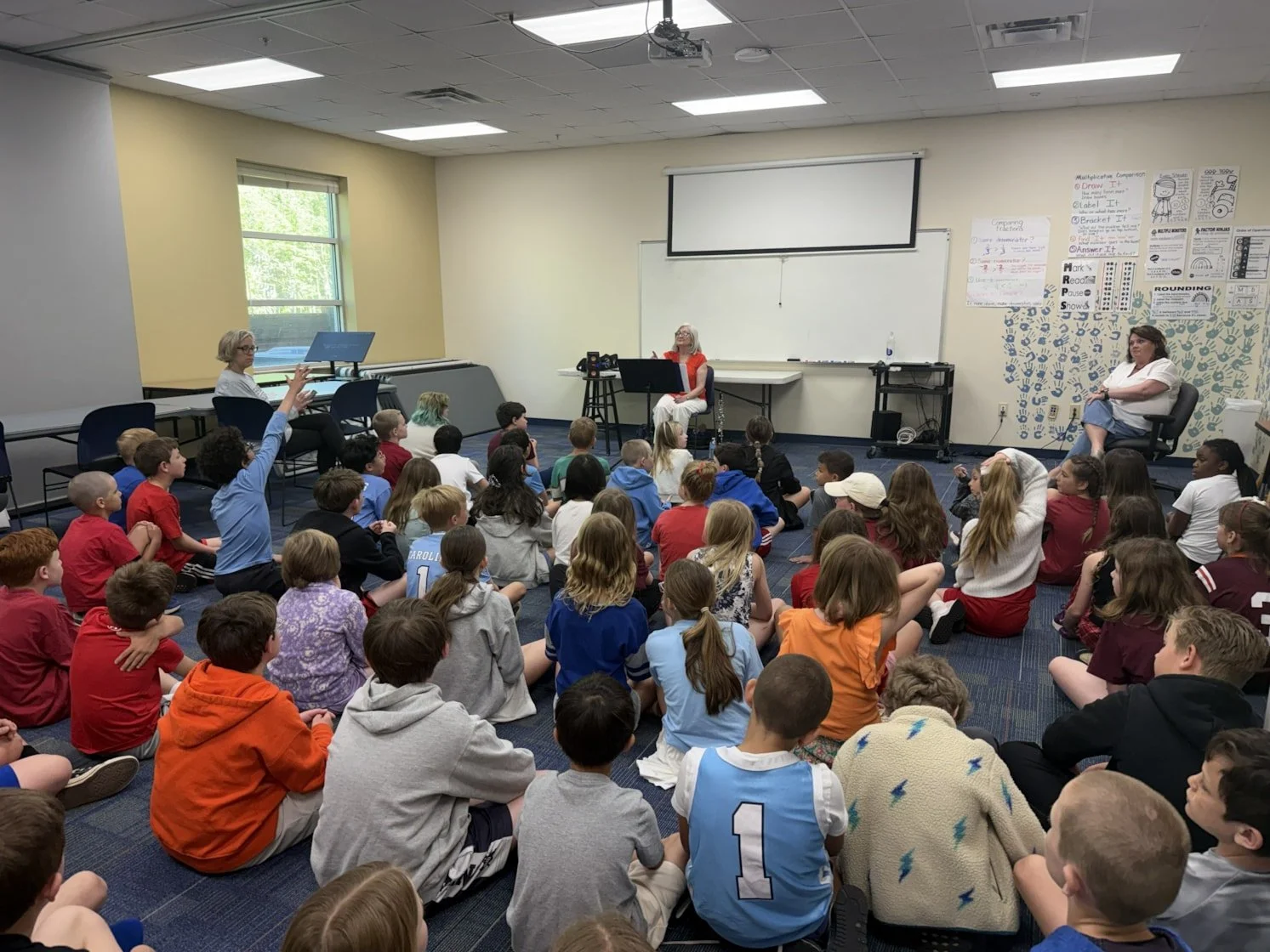 Twenty students sitting on the floor in front of an oboe player, one child is raising their hand
