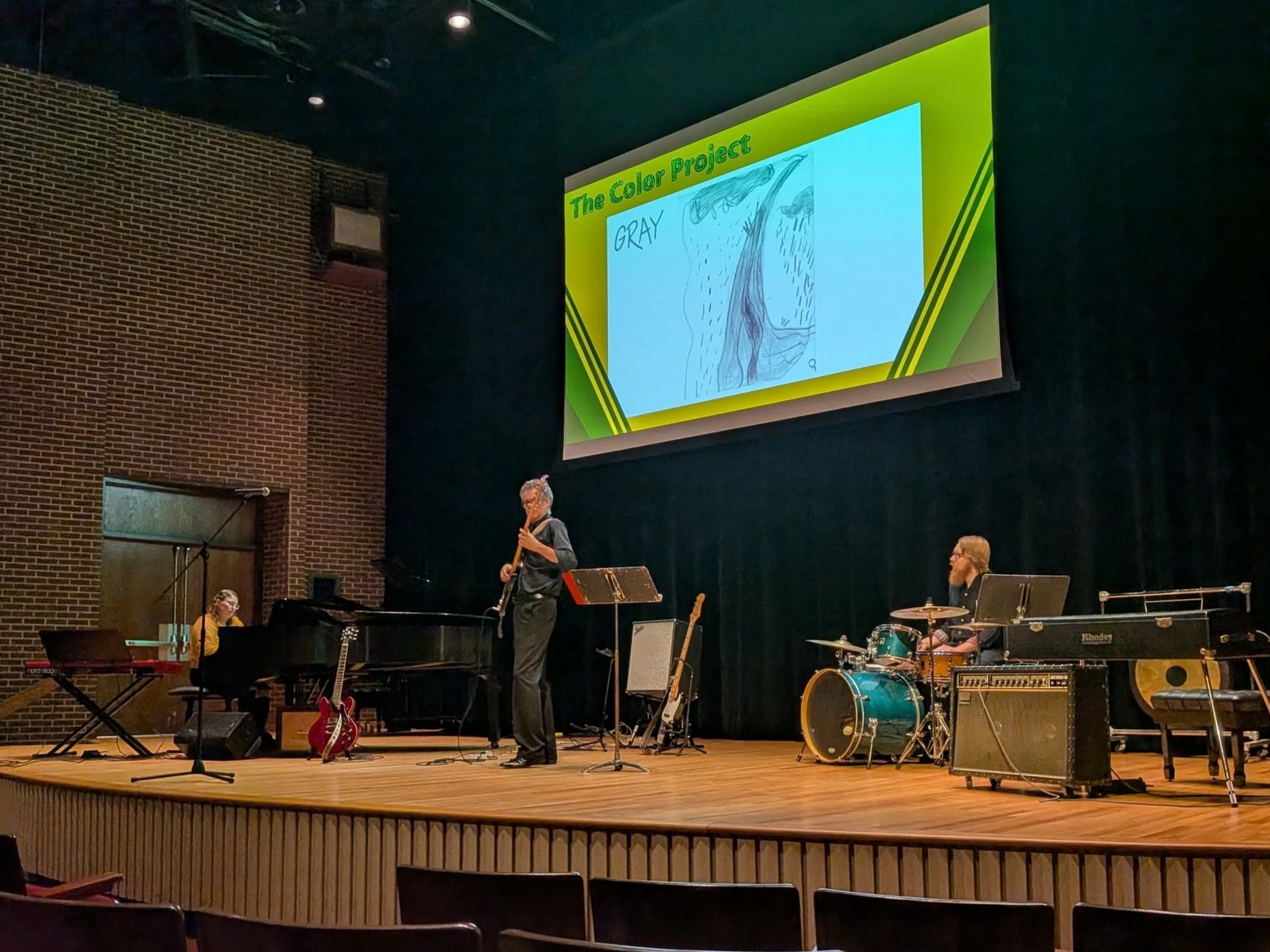 PIanist, Bassist, and Drum Set player performing under projection screen with a child's crayon drawing