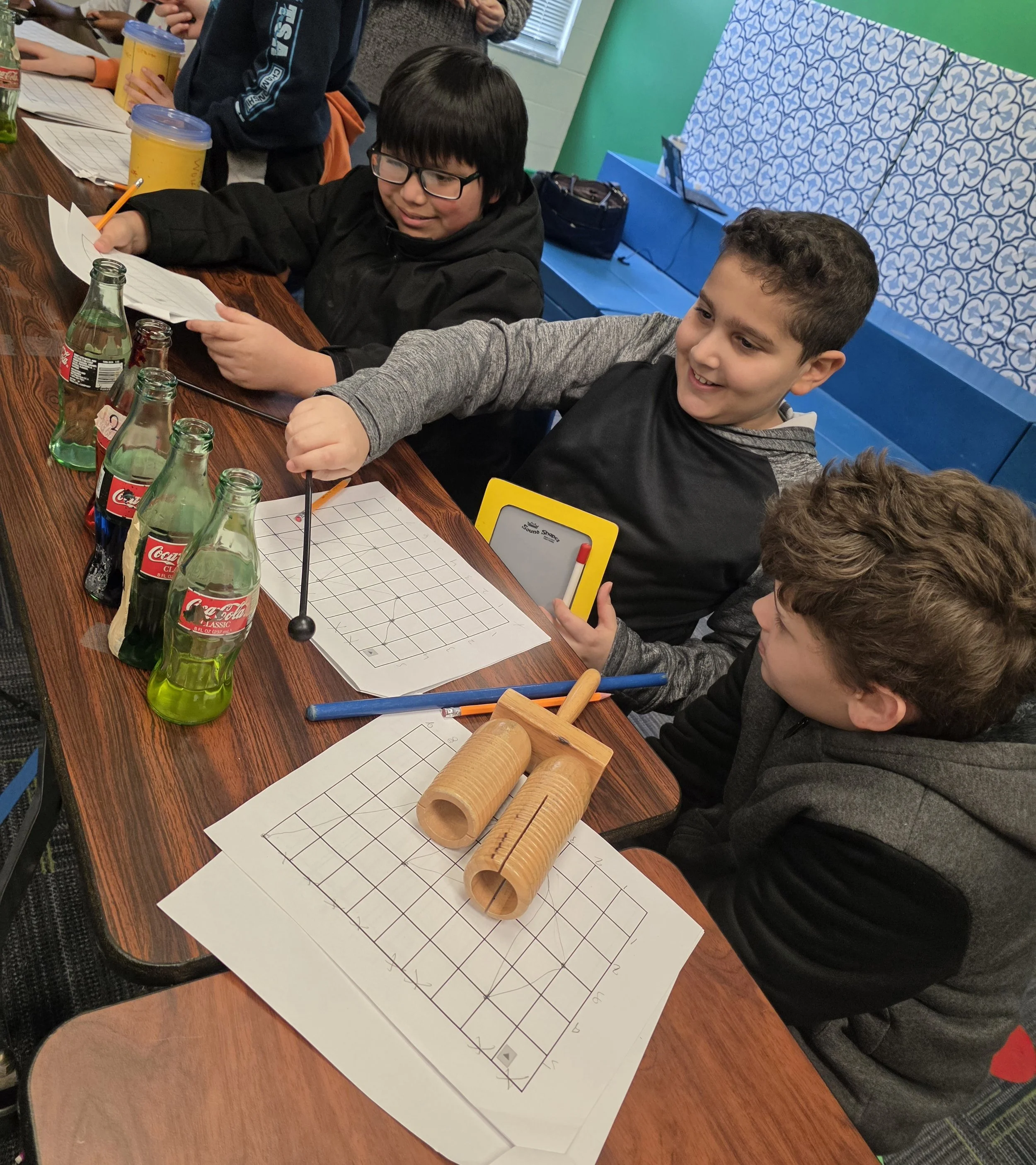 5th grade students at a table tapping Coca-Cola bottles with mallets and writing on spreadsheets