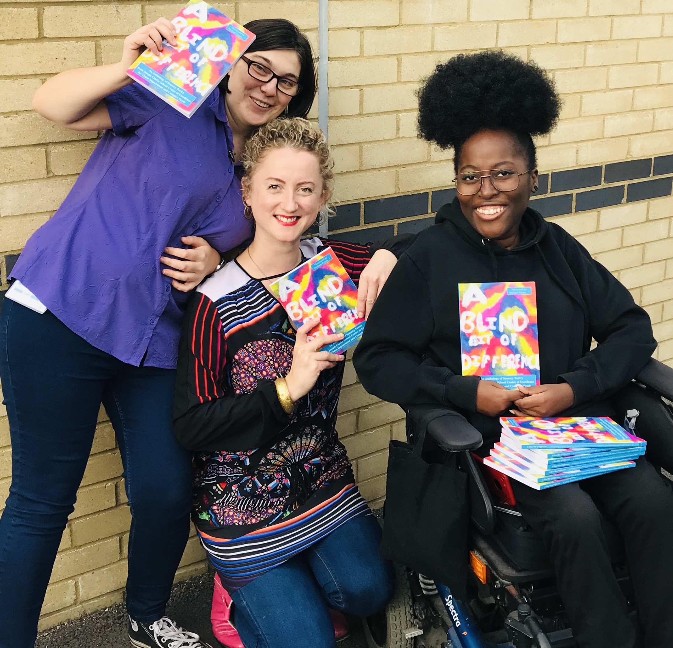 Image Description: photograph of featured artist Zara Jayne, Project manager Amy Neilson Smith and proud wheelchair user and artist Miss Jaqui pose with copies of A Blind Bit of Difference in front of a beige wall