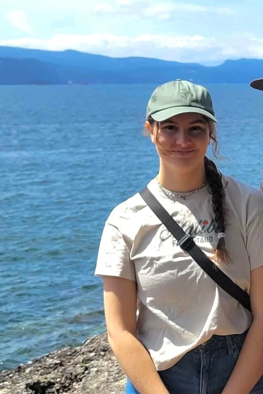 A young woman with a braid wearing a cap, t-shirt, and a crossbody bag standing by a lake with mountains in the background.