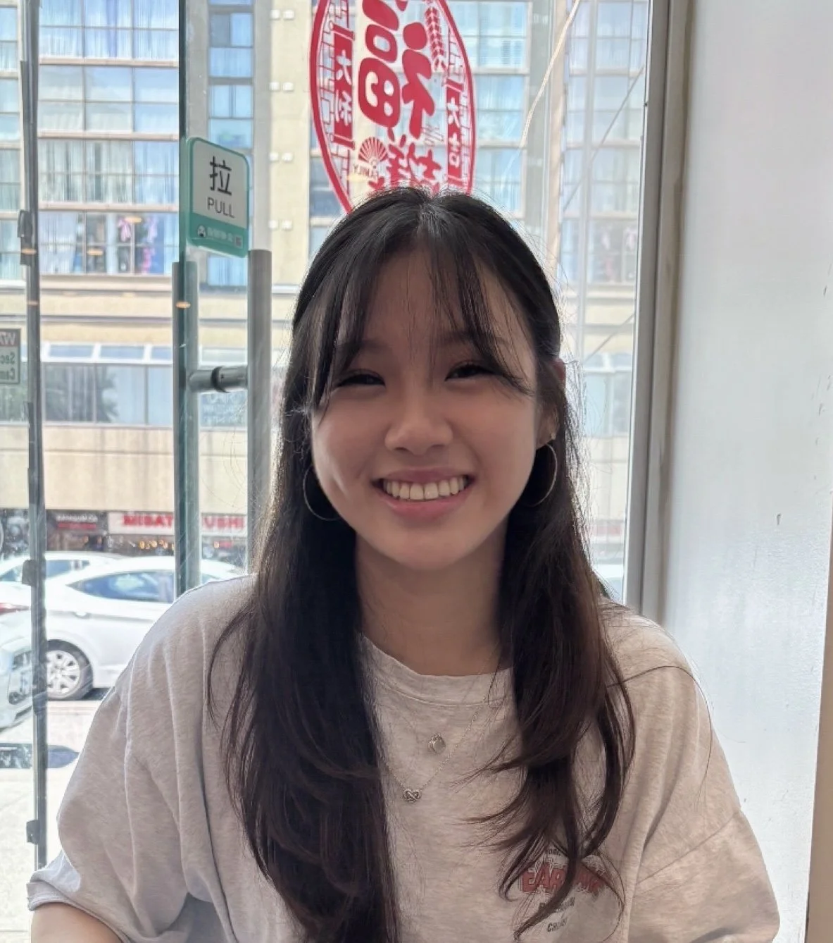A young woman with long dark hair, wearing hoop earrings and a light-colored T-shirt, smiling while sitting inside a cafe near a window.