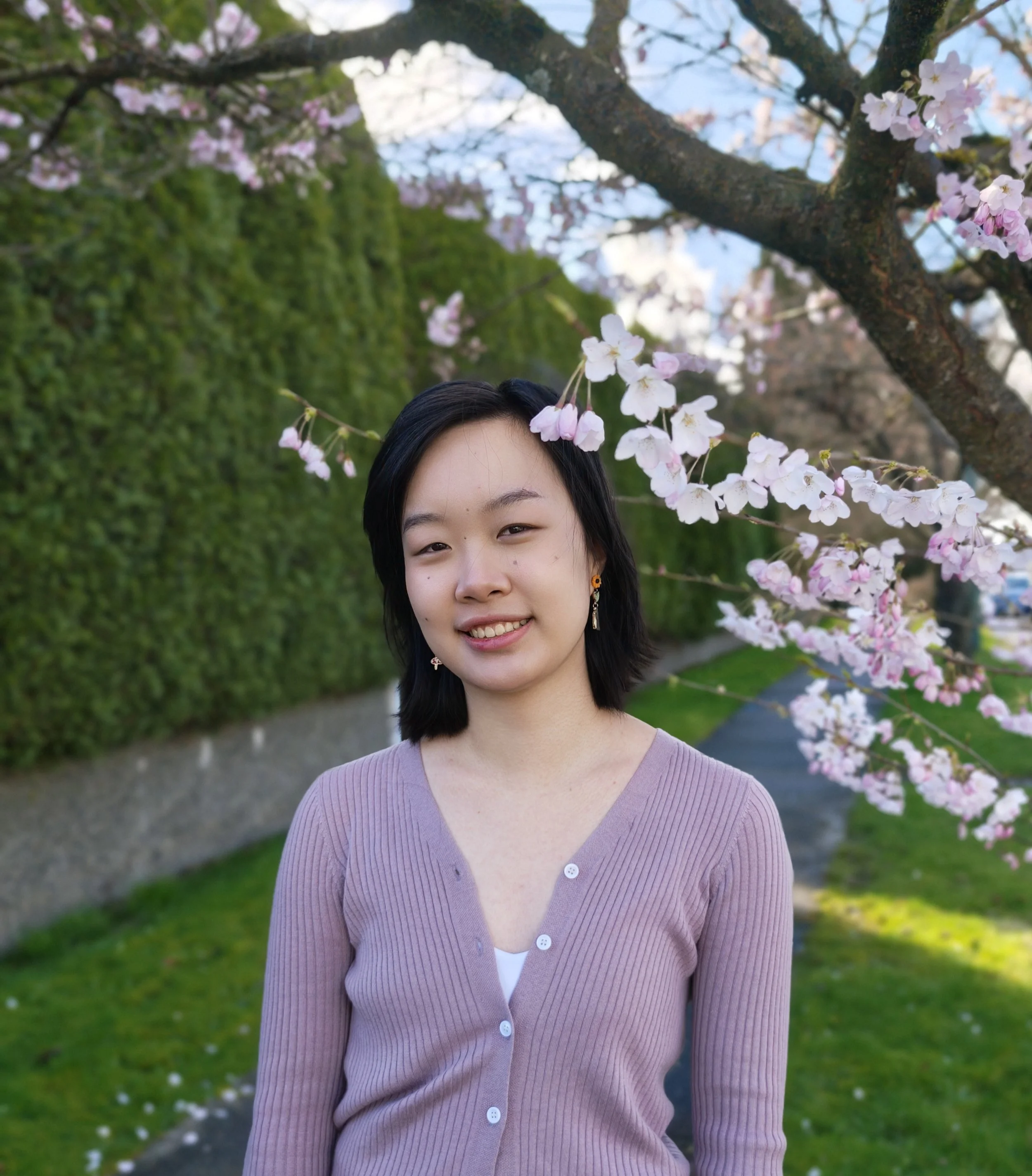 A young woman with black hair and earrings standing outdoors beneath a cherry blossom tree in bloom, with green grass and a pathway in the background.