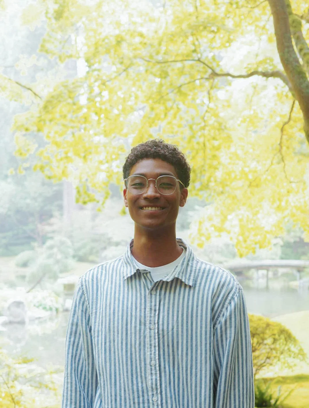 A young adult with glasses, wearing a light-colored striped shirt, smiling outdoors with yellow-green tree leaves in the background.