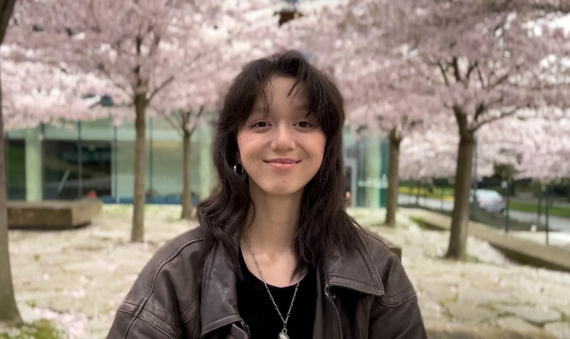 A young woman with dark hair and a silver necklace standing in front of cherry blossom trees in full bloom