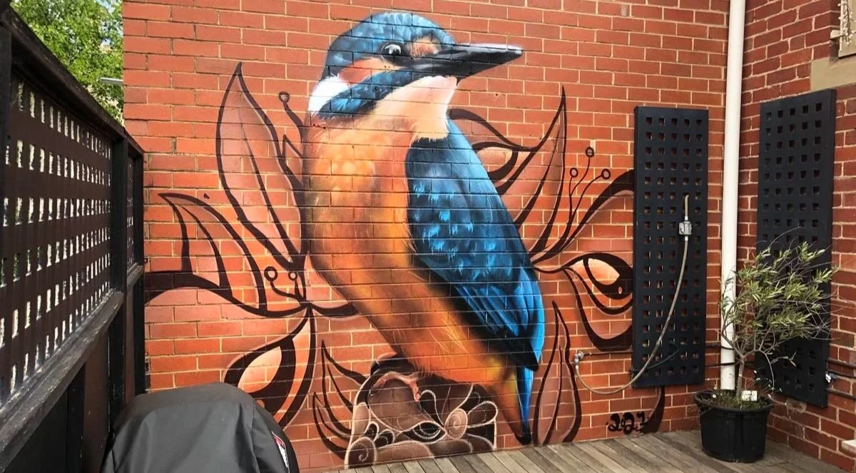 Colorful mural of a large bird, likely a kingfisher, painted on a brick wall with decorative black line art behind it, on a small outdoor patio.