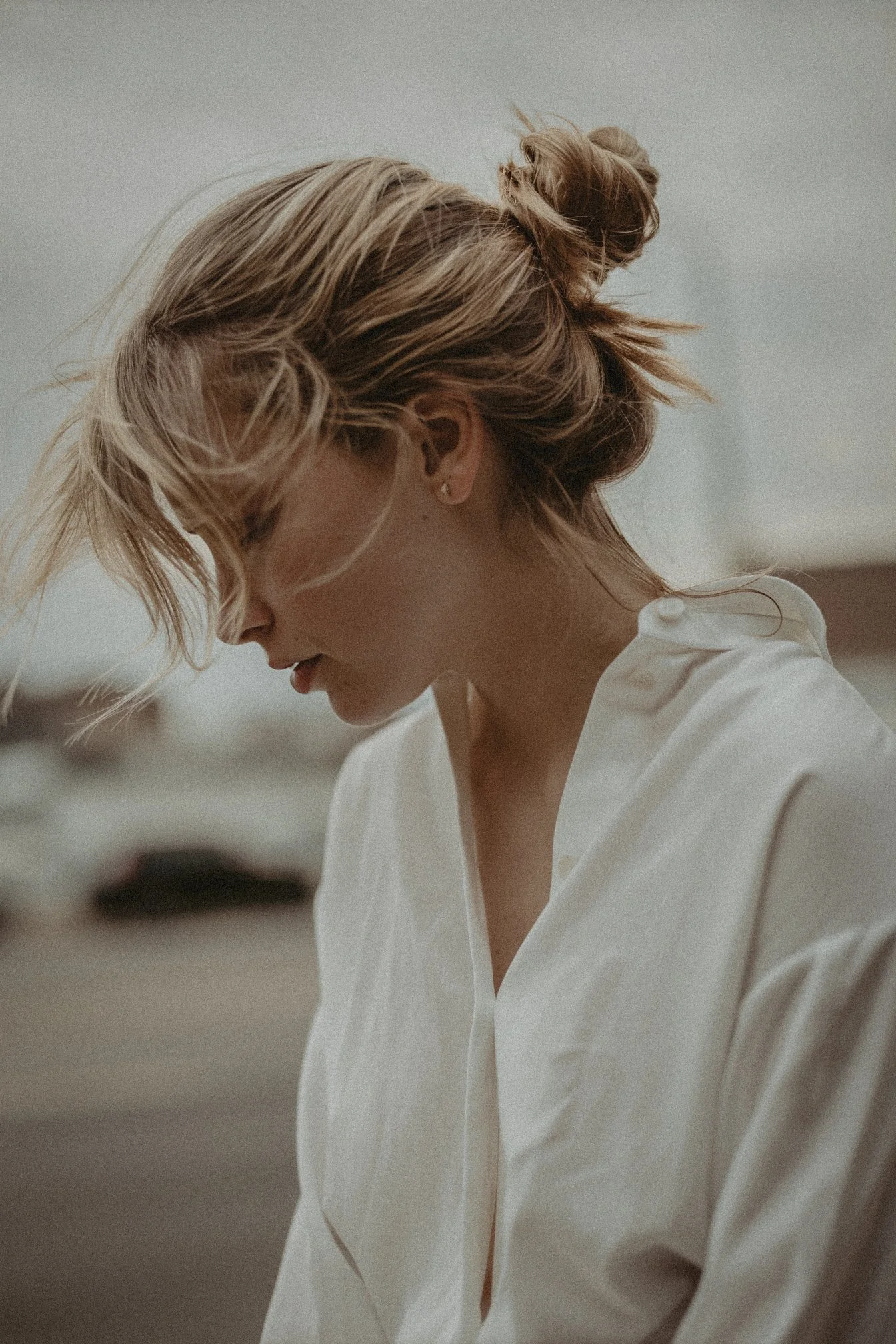 A woman with tousled blonde hair in a messy bun, wearing a white blouse, looking down with her eyes closed.