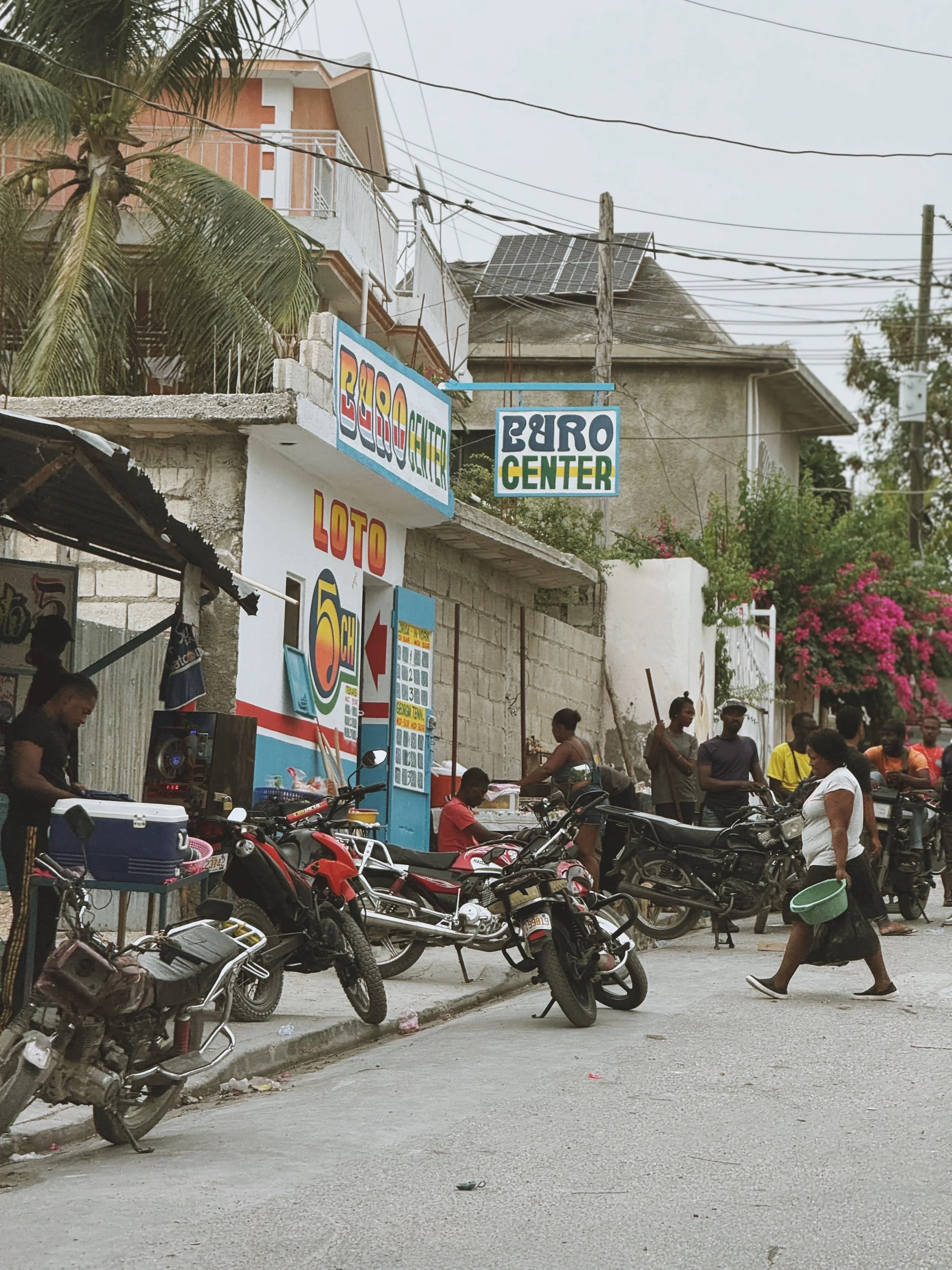 Street scene with motorcycles parked along the sidewalk, people walking, and buildings with signs and a palm tree in the background.