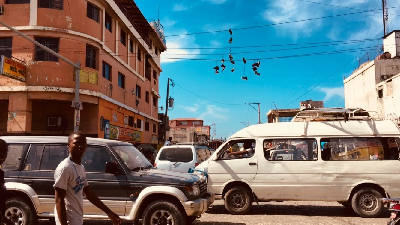 Street scene with cars and pedestrians, including a man in a white T-shirt walking. Buildings and power lines in the background, with shoes hanging from the wires and a bright blue sky.