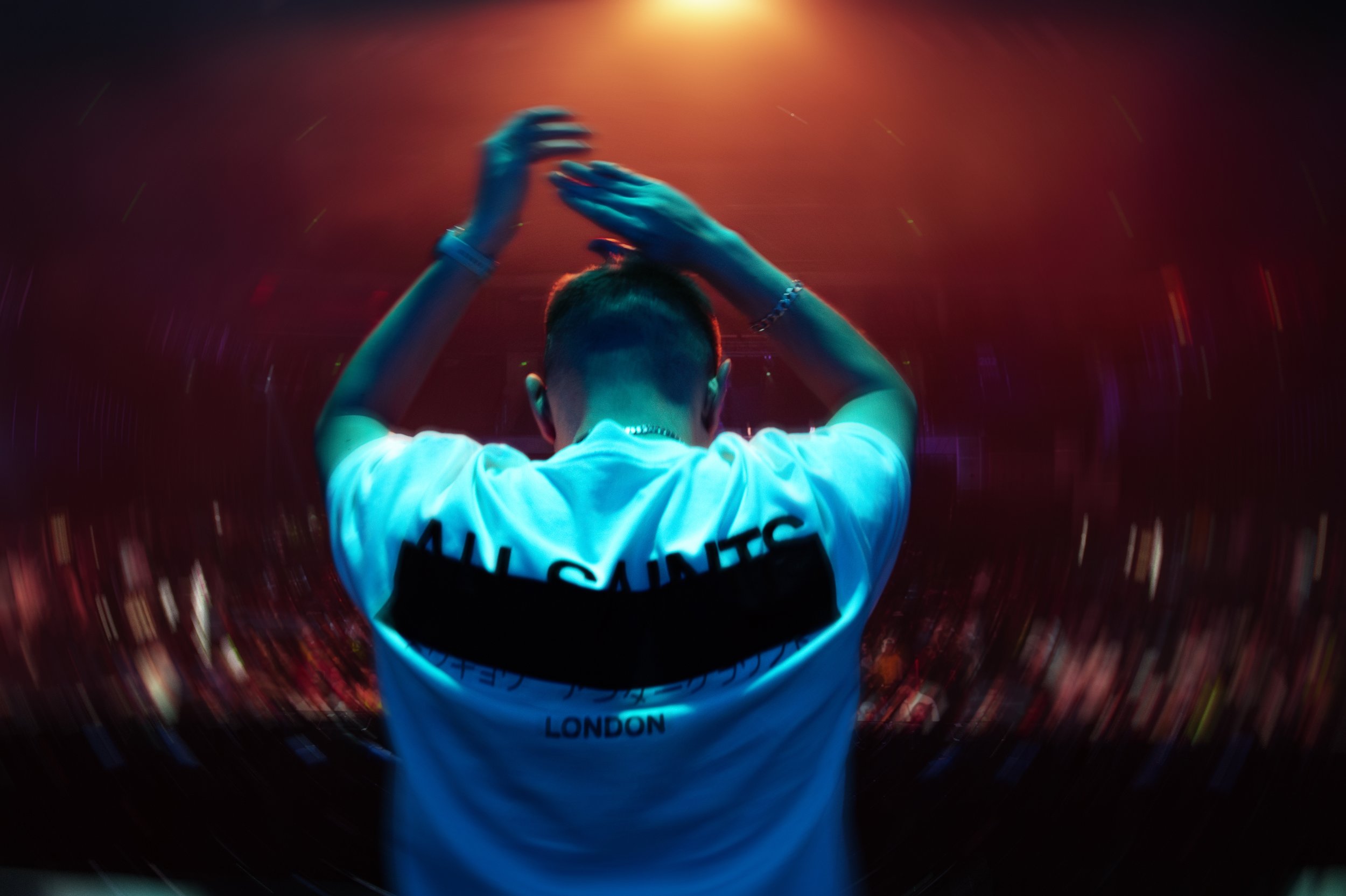 Person with short hair and jewelry dancing or performing on stage under colorful lights, viewed from behind.