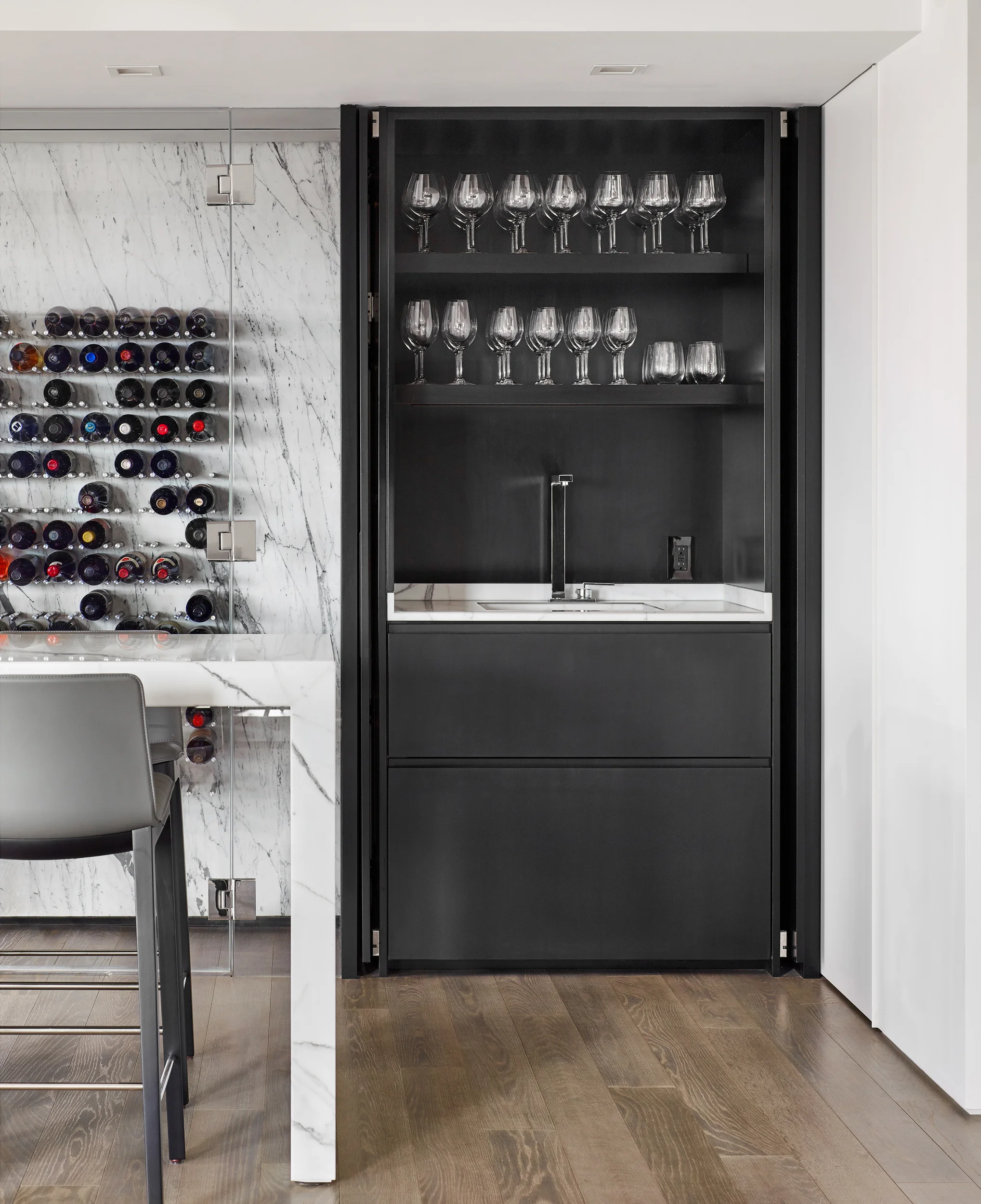 DOWNTOWN LIVING LOCATION Boston, MA COMPLETED 2018 ARCHITECT & INTERIOR DESIGN Walker Architects GENERAL CONTRACTOR The LaGasse Group STONE SUPPLIER & FABRICATOR Cumar PHOTOGRAPHER Jared Kuzia Photography Our clients, downtown academics with grown children, sought to combine two mid-rise apartments to gain an unparalleled elevated view sweeping from the dome of the Boston State House westward to the riverfront campuses of MIT and Harvard. Because the owners were already comfortably at home in one of the apartments, the added second unit was designed as additional spaces for relaxation, with accommodations for billiards and darts, a seating area focused on a dramatic new fireplace, a sophisticated wine bar, and an adjacent study with sleek guest bath. New finishes were extended throughout the “public” areas of the combined apartment, and include a newly paneled entry hall with concealed closets and a vegetated wall facing the front door. In a home where the view is always going to be the star, new spaces are united in their modern simplicity, careful detailing and luxurious choices in materials. Anchoring a 45-foot long living room, a plane of warm brown marble is incised with a matte black steel fireplace that mimics the horizontal glow of the room’s daily western sunsets. Nearby, wine is served on a white marble pattern-matched table, backed by a display of bottles in a conditioned “cellar” lined with the same marble cut thin and backlit. Flanking the wine storage and display are paired dark bolivar cabinets housing serveries with concealed refrigeration and dishwashers. The guest bath, a study in smooth grey and white, features frameless glass, concealed ribbons of light and super-minimalist washbasins carved into a floating quartzite ledge. All spaces are wrapped in crisp white plaster without trim or baseboards, floating above a new floor of richly stained oak.