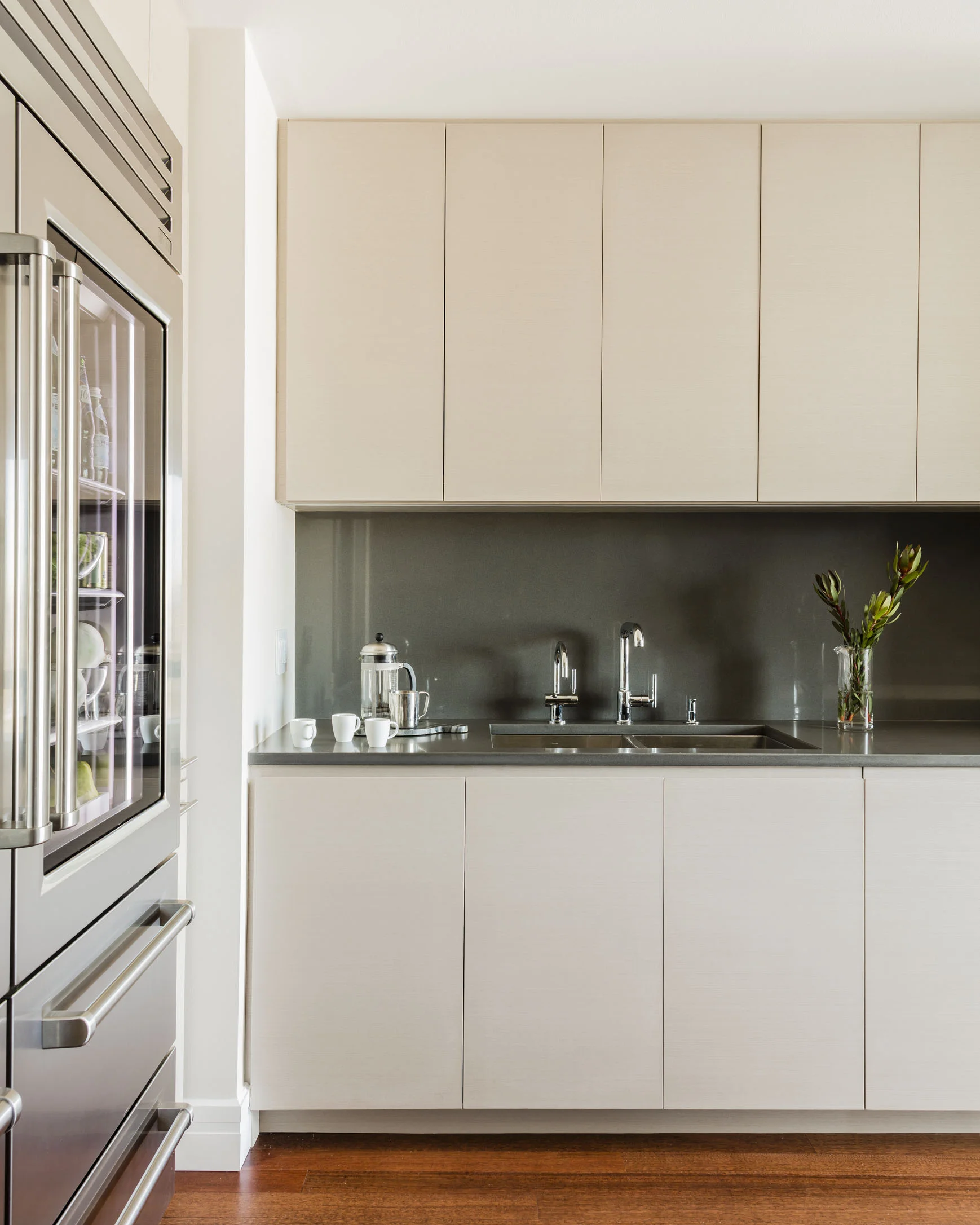 Modern kitchen with beige cabinets, stainless steel sink, and dark gray backsplash. White cups are on the counter, a glass vase with green and red plants is on the right, and a stainless steel refrigerator is on the left.