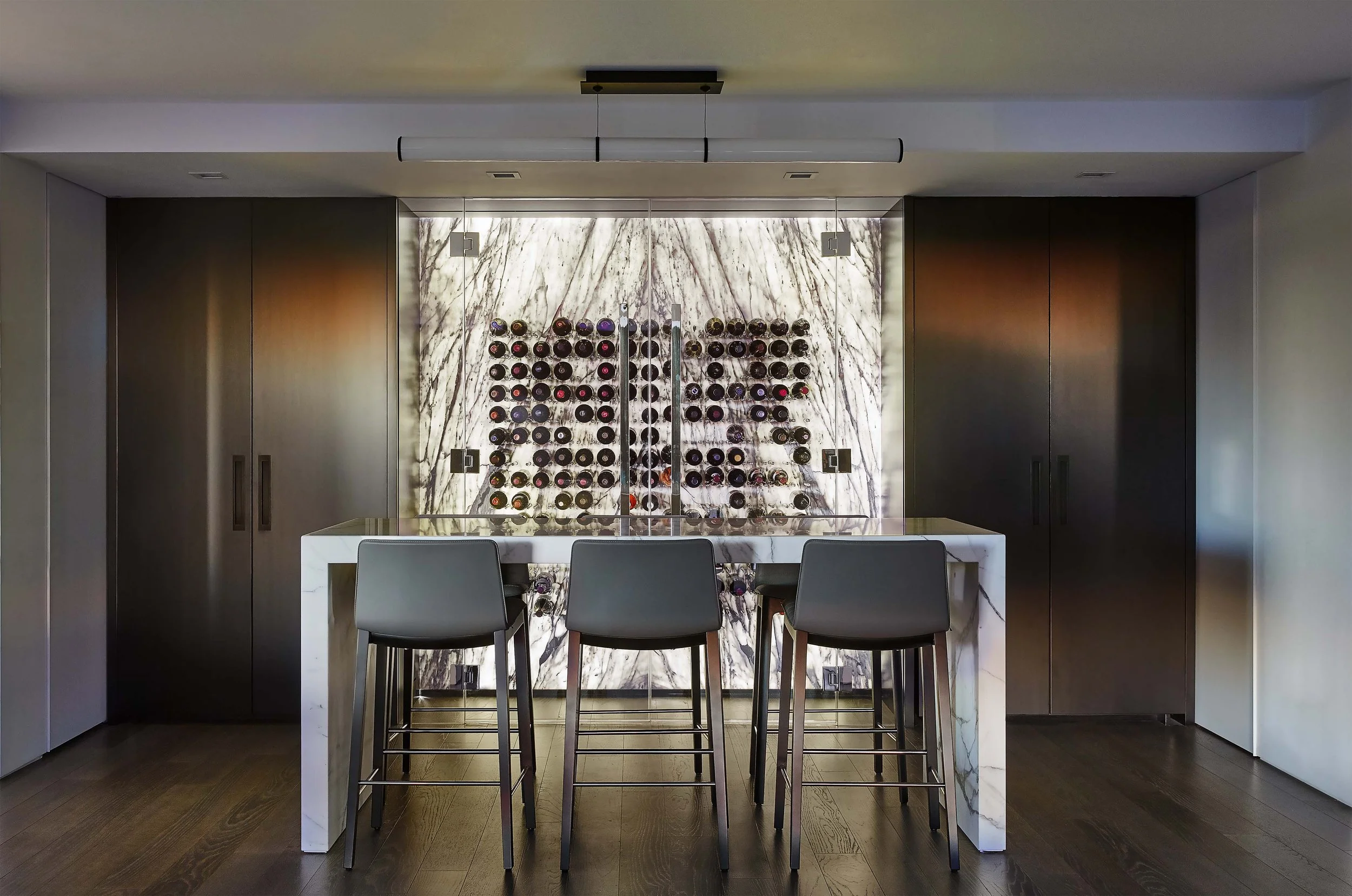 A modern home bar finished in dark wood cabinetry and featuring backlit marble wine storage
