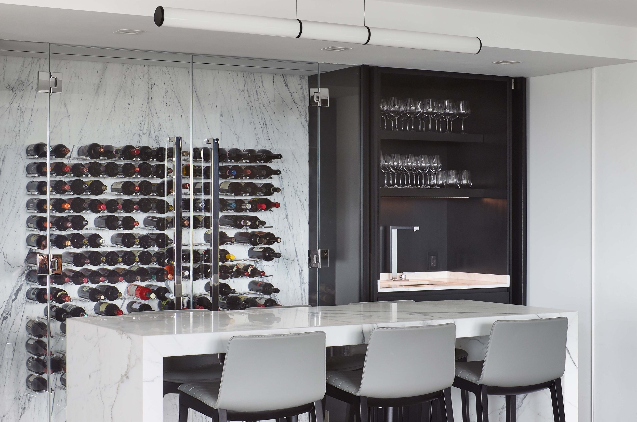 A modern wine storage area with a glass-front wine rack holding multiple bottles and a black cabinet with glassware on shelves. A white marble counter is in foreground with white chairs around it.