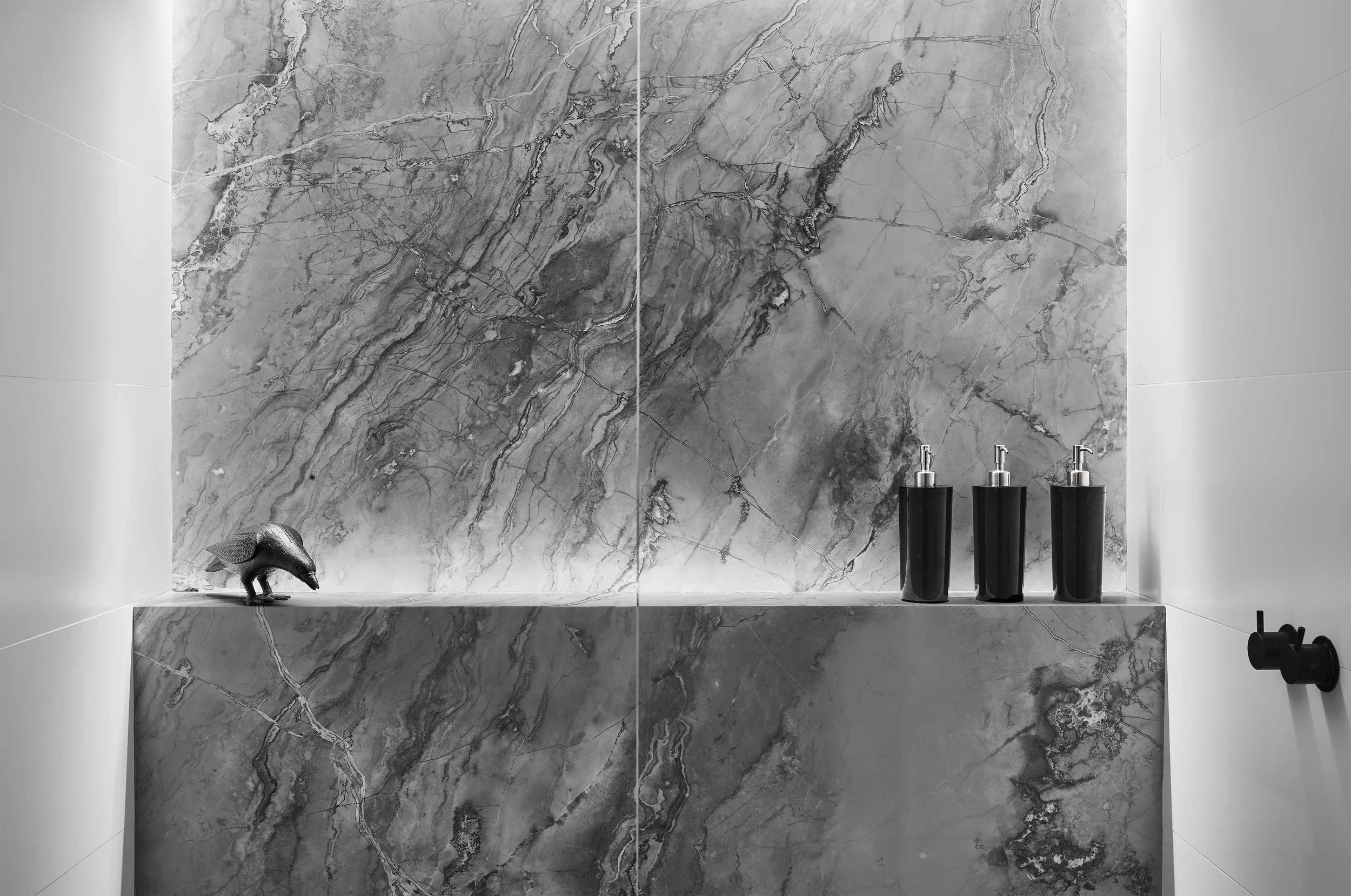A bathroom shelf with a dark stone sculpture of a pig, three black bottles with silver pumps, and a black long object against a large grey marble wall with white veining.