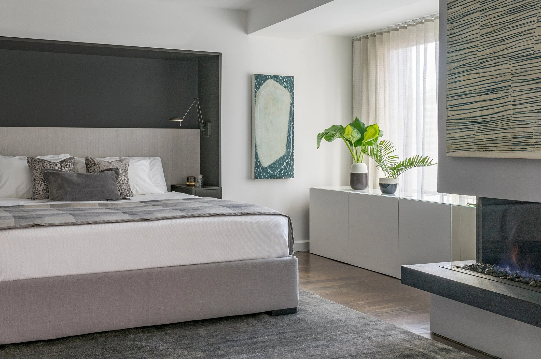A primary bedroom in a renovated condominium featuring a built-in architectural headboard and a three sided gas fireplace
