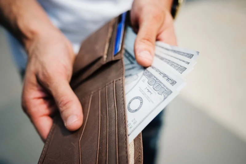 Person holding a brown leather wallet containing dollar bills.