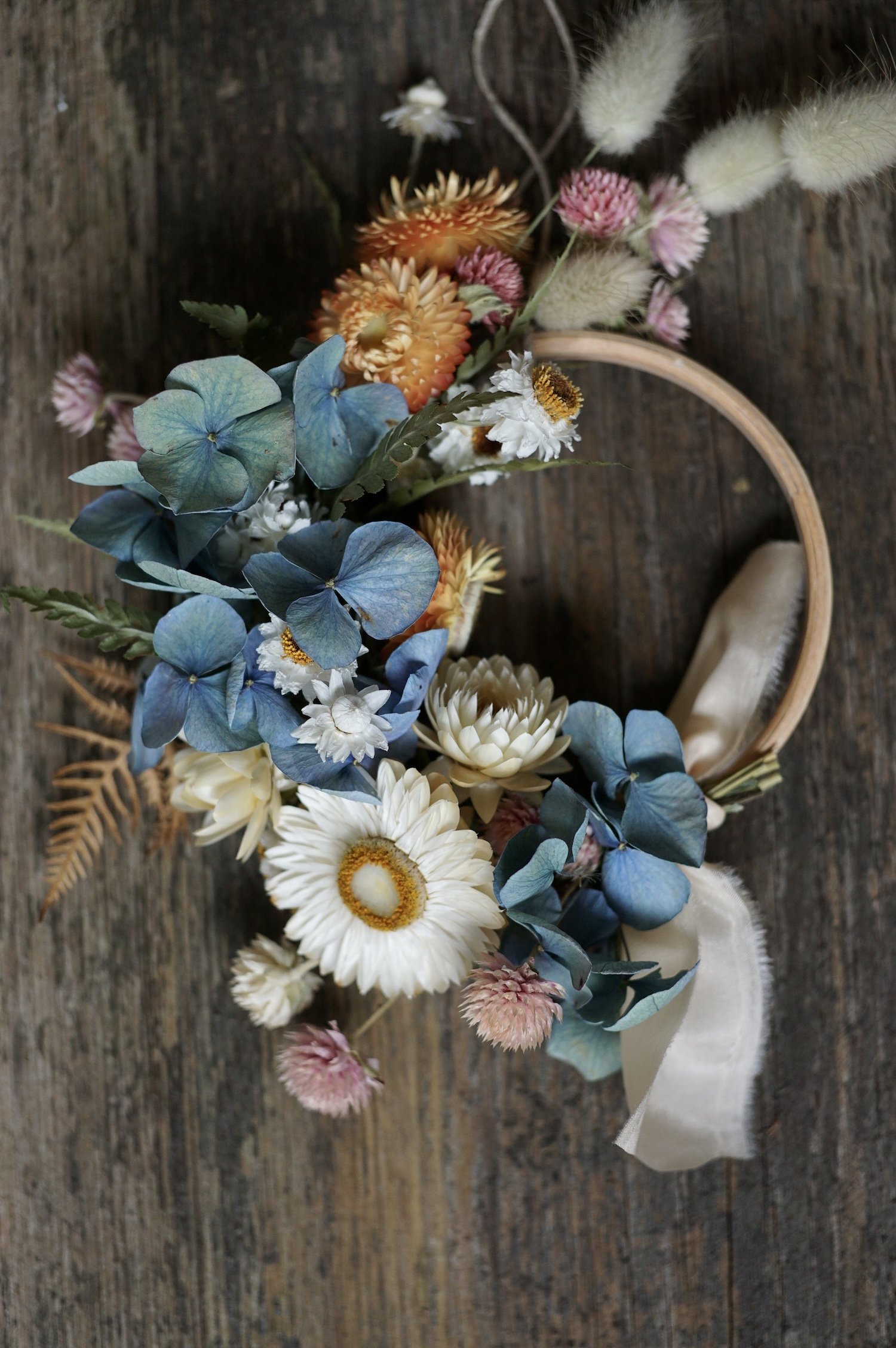 Mini dried flower wreath of pastel coloured flowers and a pale peach ribbon on a wooden surface.