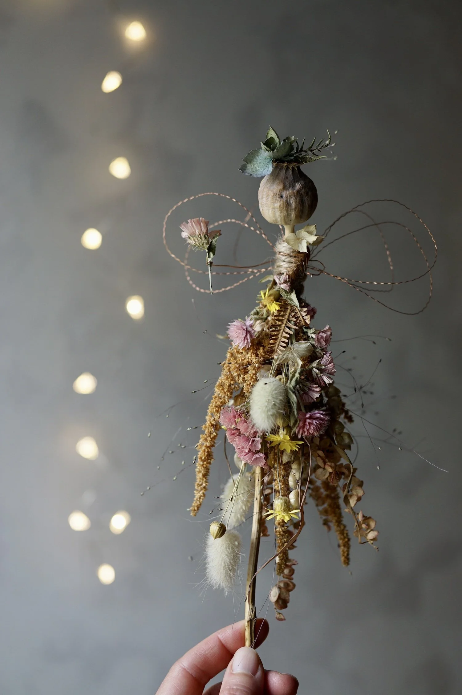 Flower fairy made with a poppy seed head and dried flowers with wire for wings and arms, near fairy lights.