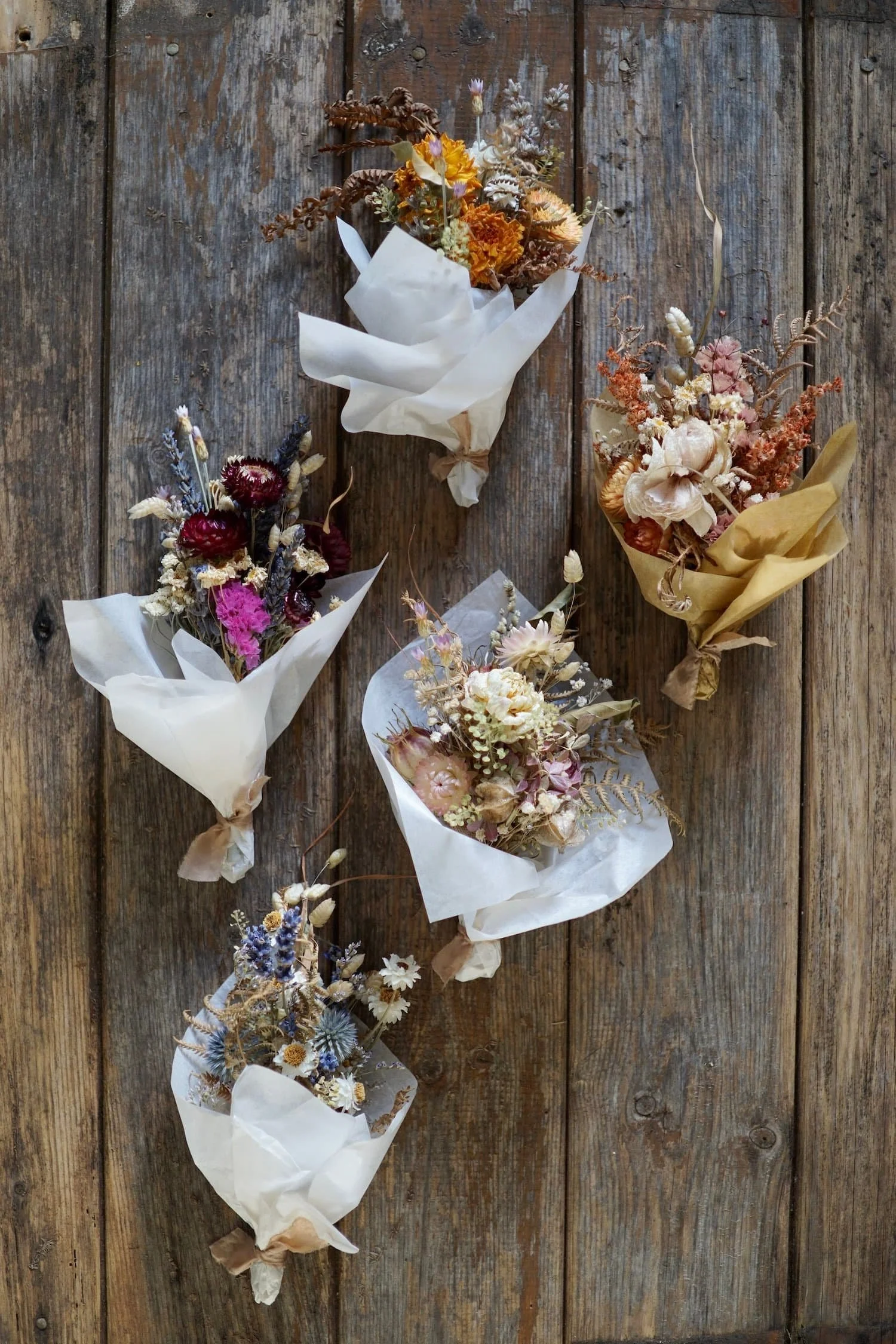 Five small dried flower bouquets in different colours, wrapped in white and brown tissue paper and tied with ribbon, lying on a weathered wooden surface.