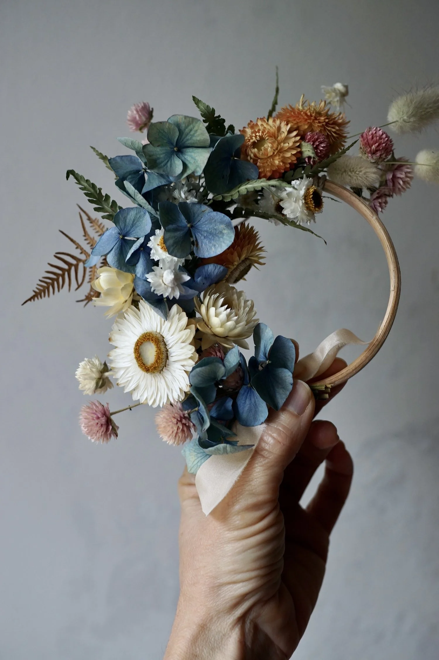 Mini dried flower wreath of pastel coloured flowers and a pale peach ribbon held by a hand.