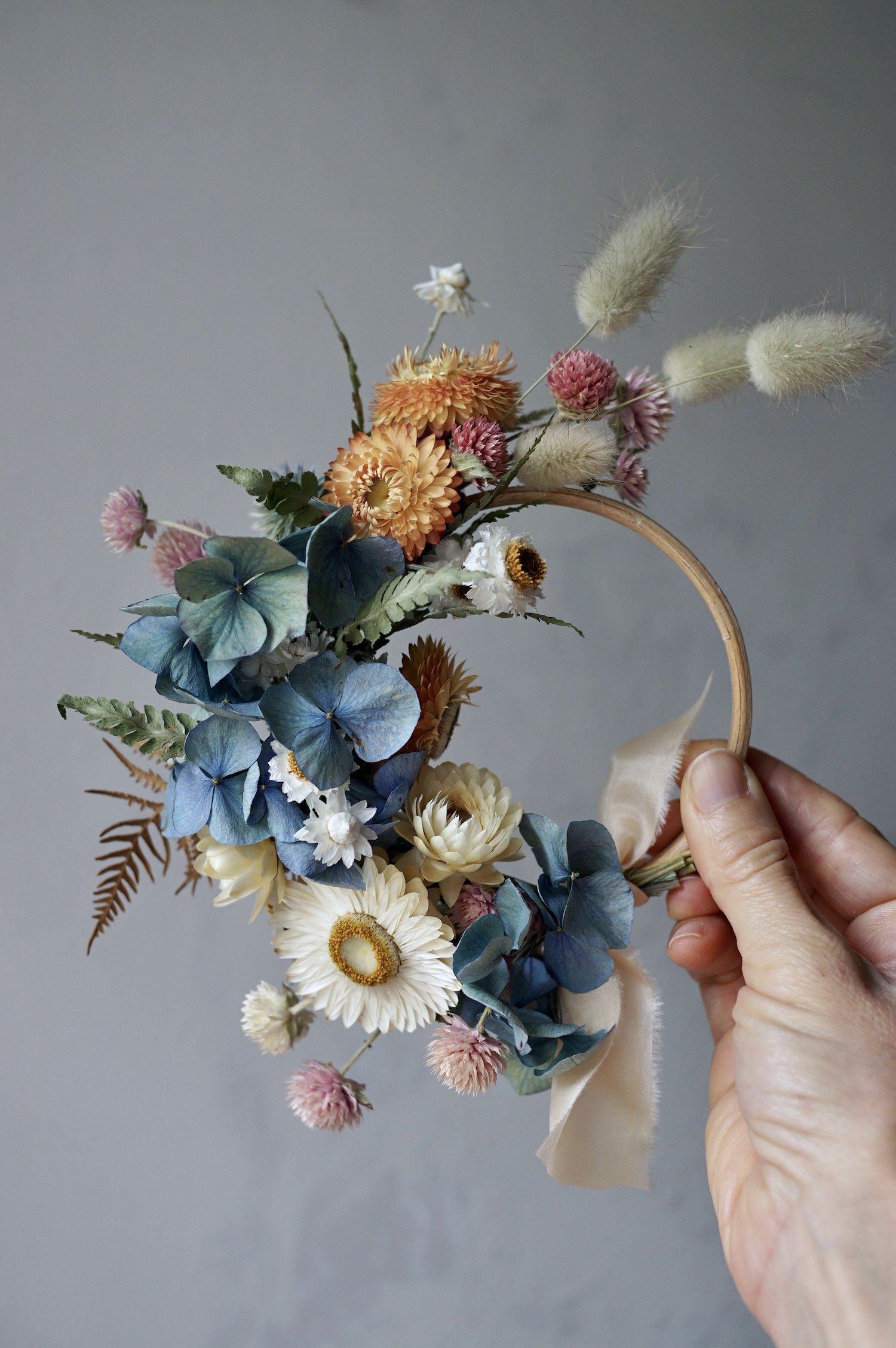 Mini dried flower wreath of pastel coloured flowers and a pale peach ribbon held by a hand.