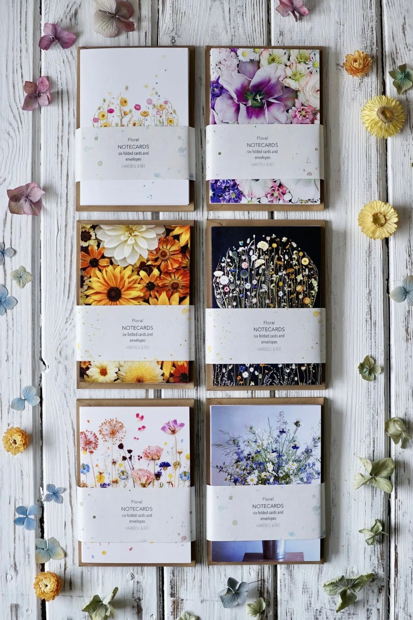 Six packets of notecards of pictures of different flower designs lay next to each other on a white painted wooden surface, surrounded by dried flowers of various types and colours.