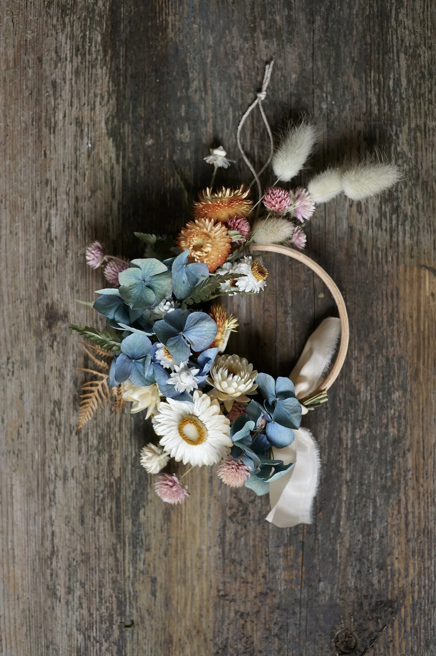 Mini dried flower wreath of pastel coloured flowers and a pale peach ribbon on a wooden surface.