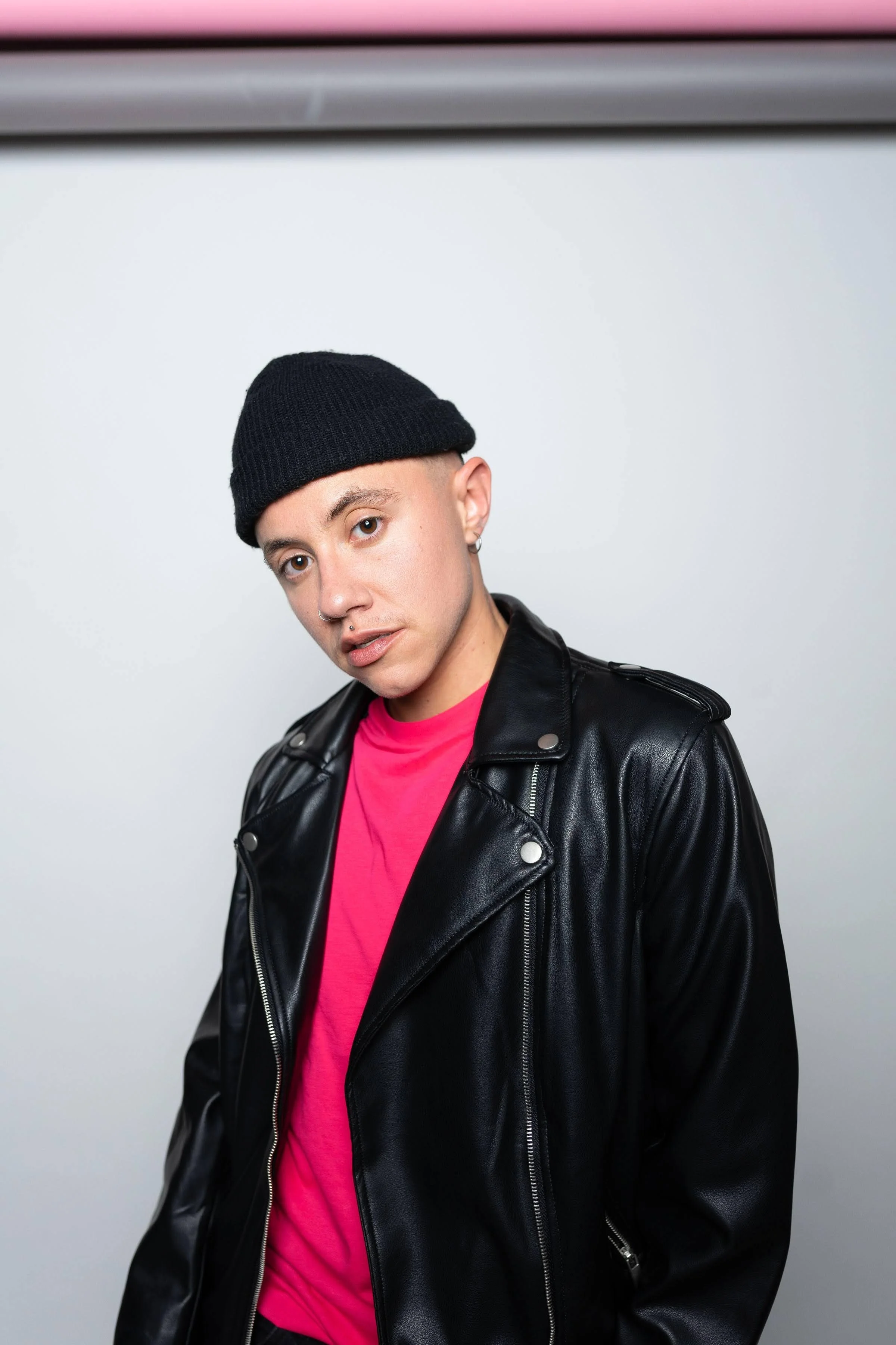 Canyon is wearing a black leather jacket over a red Tshirt, and a black beanie. They are looking at the camera, standing against a white wall.