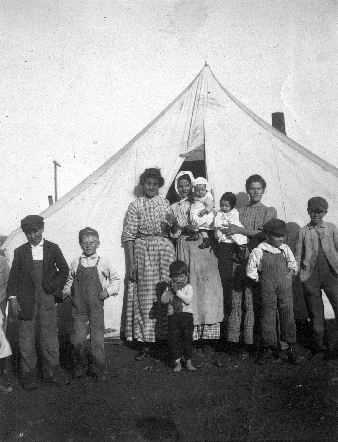 Black and white photograph of a working-class family with young children standing in front of a large canvas tent at the Ludlow Colorado coal miners' strike camp, circa 1913–1914.