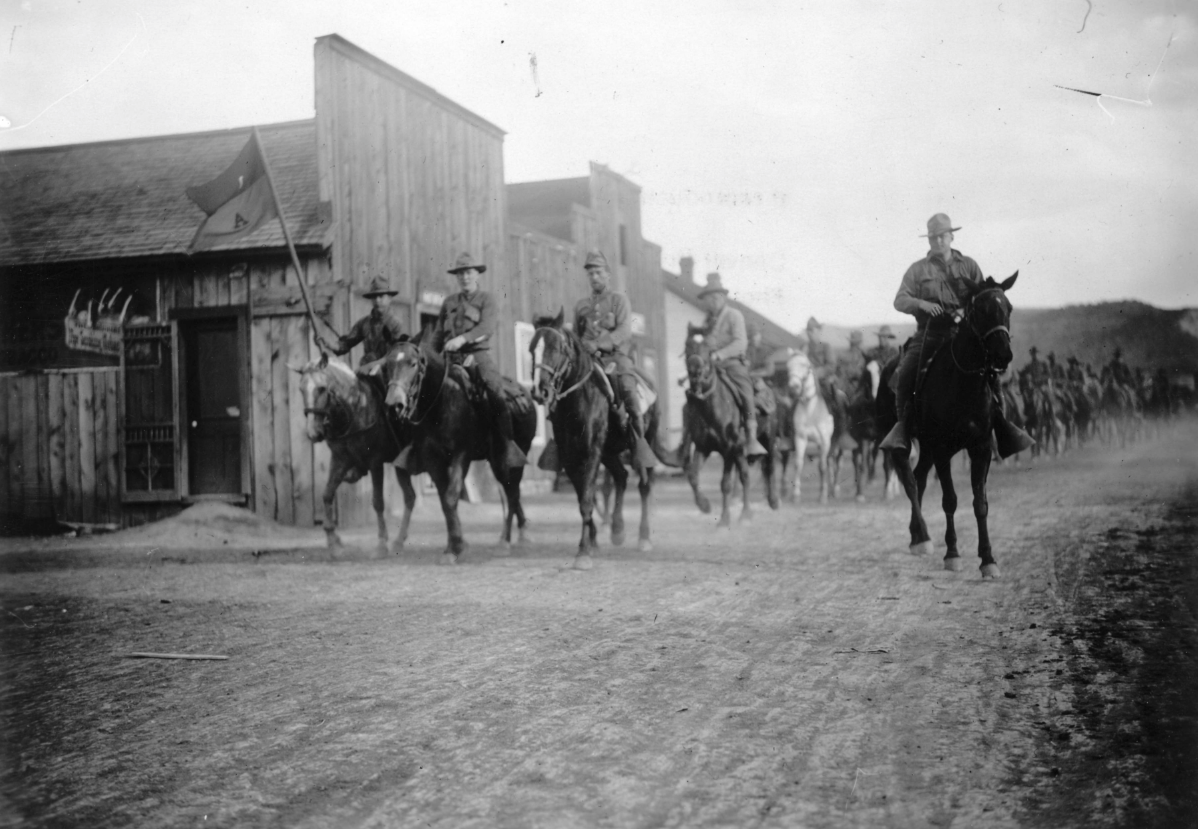 Black and white photograph of a column of armed cavalry soldiers on horseback riding down a dirt main street lined with wooden storefronts in a small Colorado town during the 1914 Coalfield War.