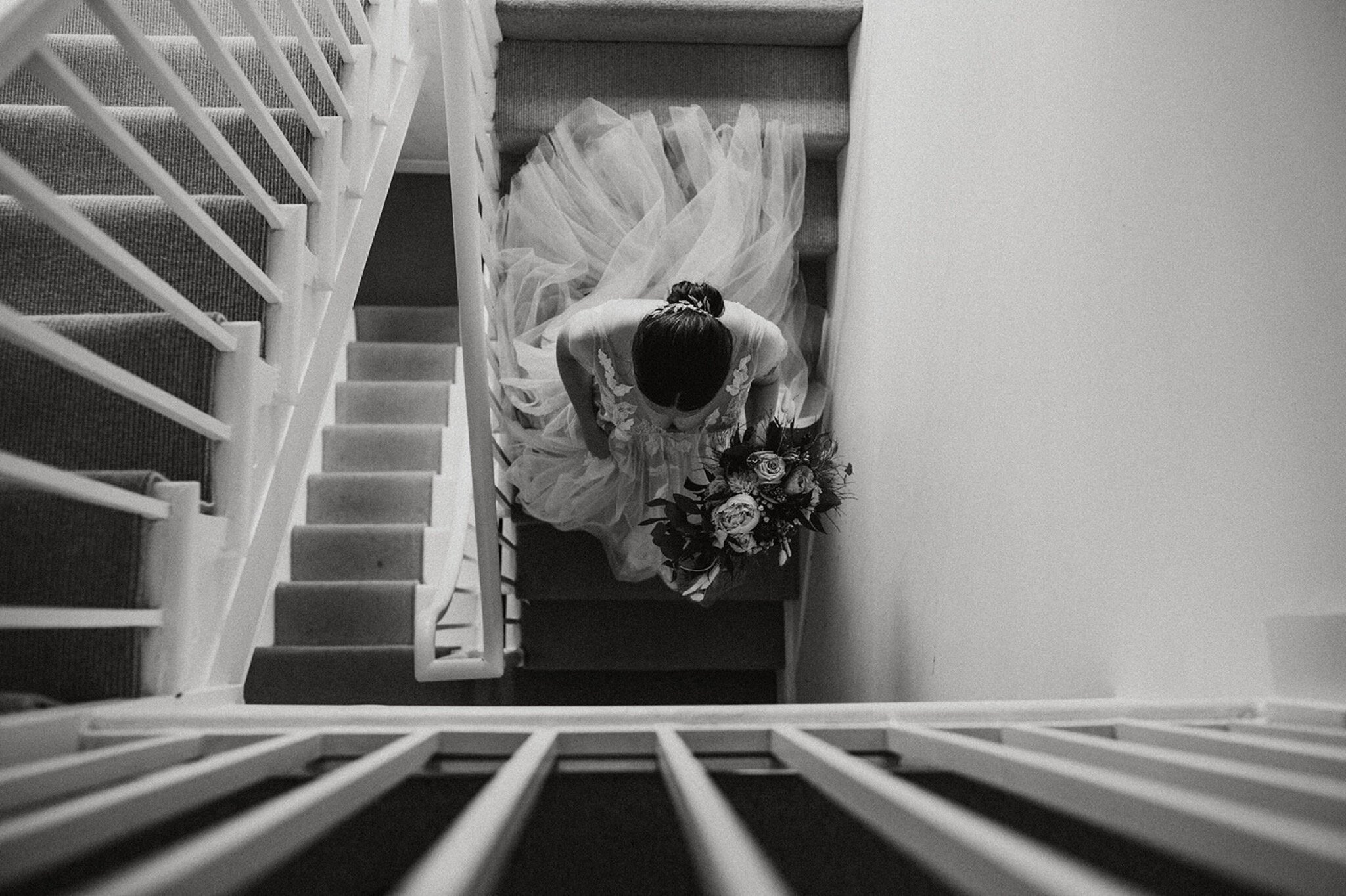 10_V+D Wedding-123_Black and white photo shot from above of a bride descending the stairs in her wedding dress with a bouquet.%0A.jpg