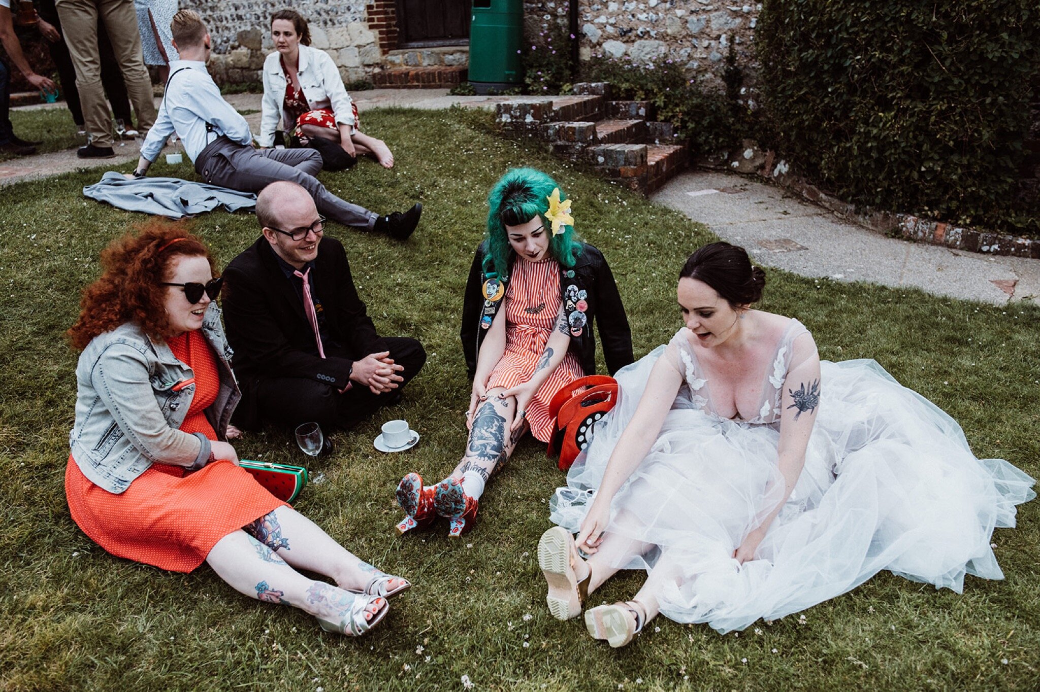 46_V+D Wedding-746_bride and guests sat on grass looking at brides shoes%0A.jpg
