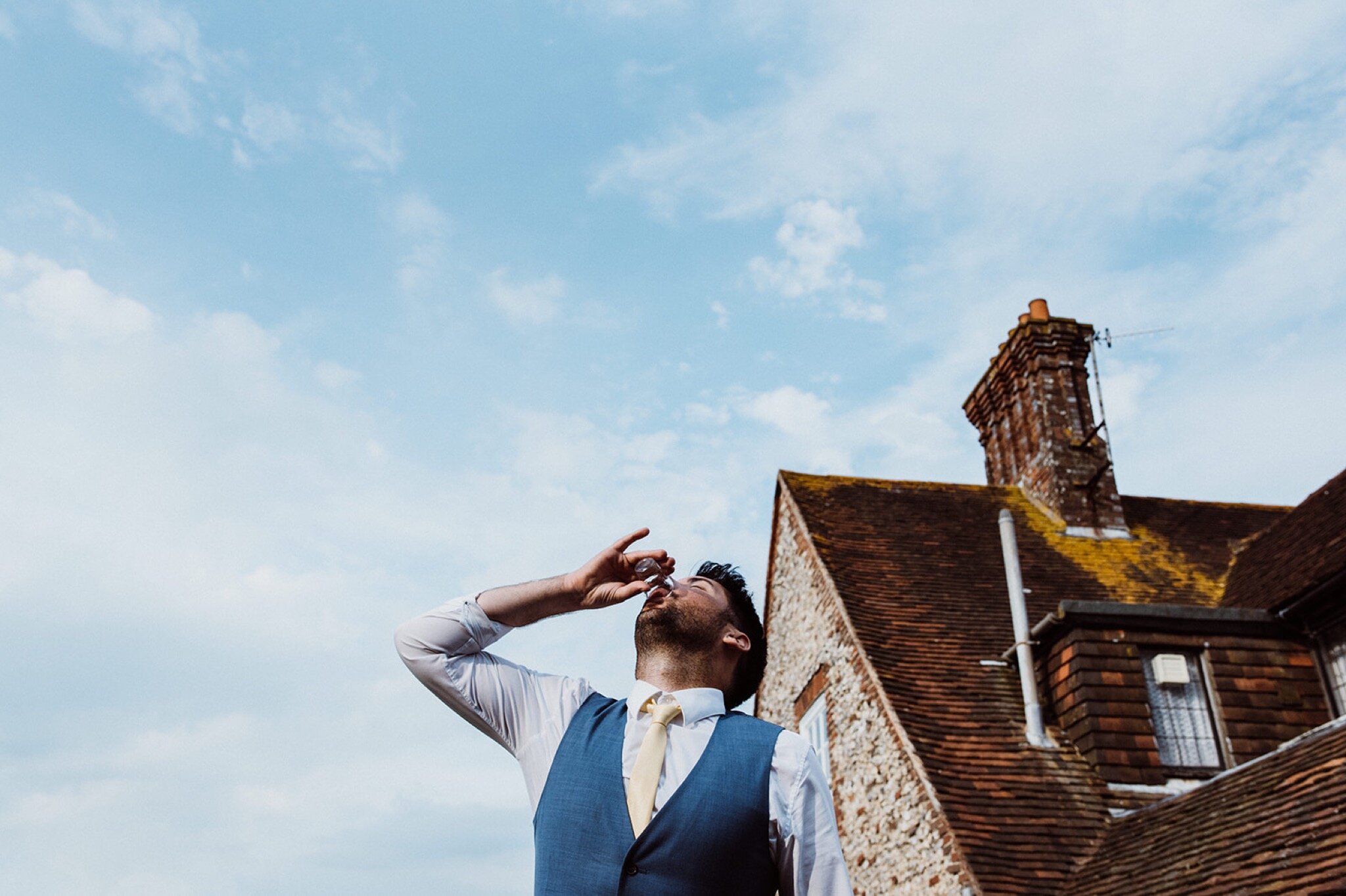 49_V+D Wedding-804_male guest in waistcoat and tie doing a tequila shot agains a blue sky%0A.jpg