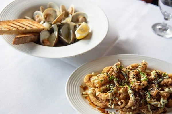 A plate of fried calamari garnished with black sesame seeds and chopped herbs, with a side of toasted bread and a basket of clams with lemon wedge.