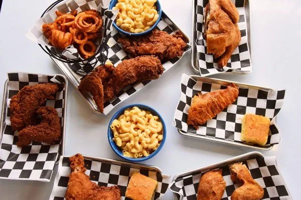 Assorted fried chicken pieces, macaroni and cheese, and onion rings served on metal trays lined with checkered paper.