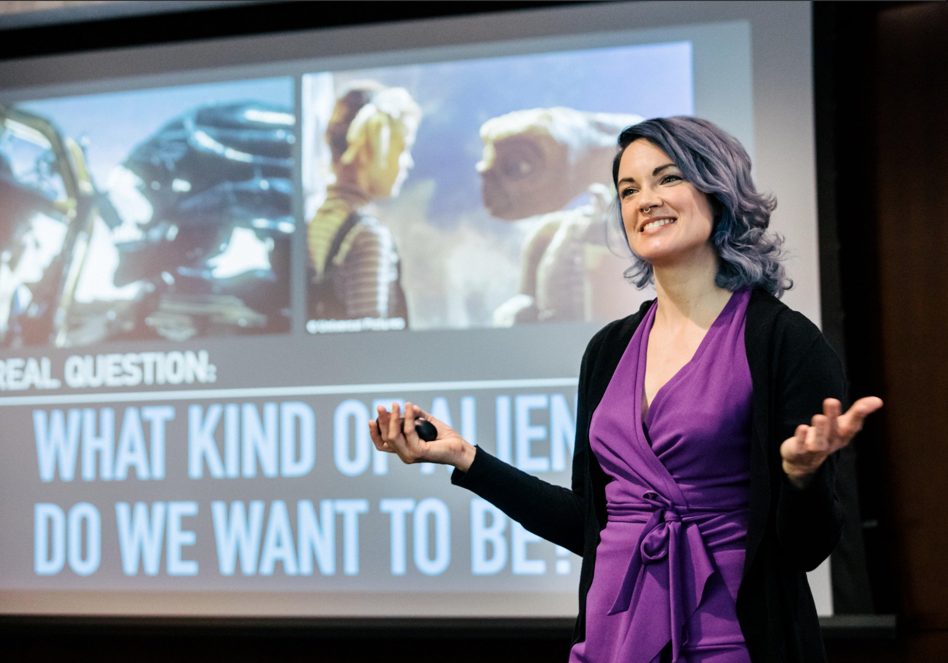 An action shot of Lucianne Walkowicz, smiling on stage in front of a slideshow
