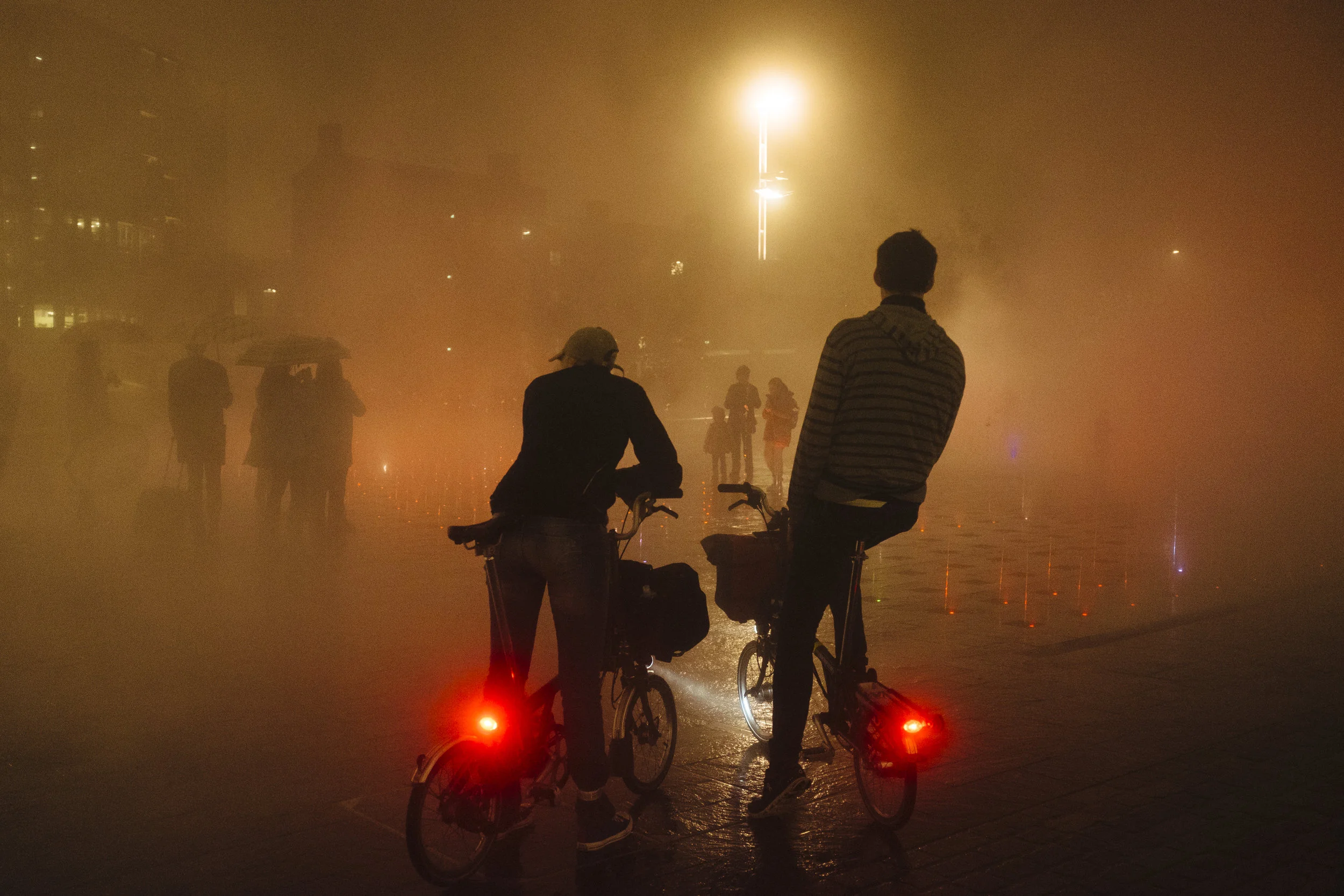 Men on Brompton bicycles look at people walking through the mist rising from the fountains at Granary Square, London, England, Saturday, Nov. 12, 2016.