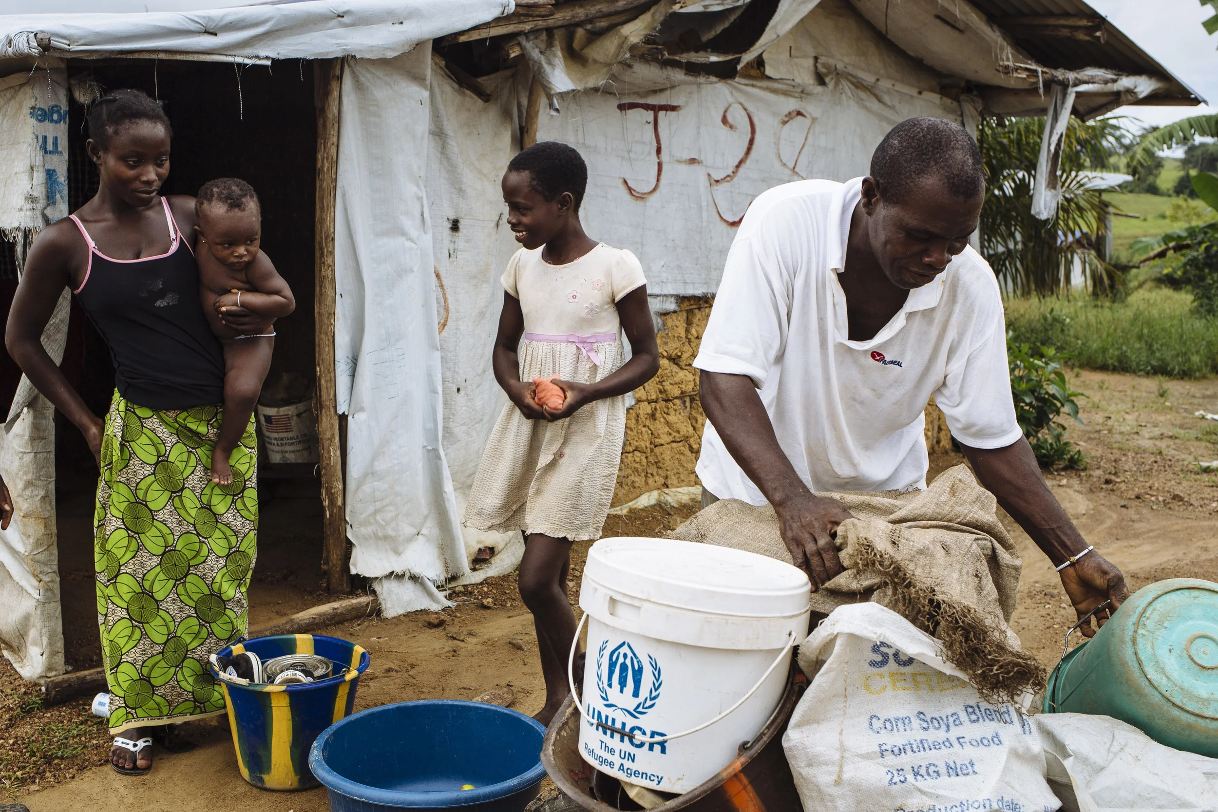 Ivorian refugees Yagou Gbordi Hie, left, holds her daughter Gnepadi Christine, alongside her sister Tahadi Hie, center, as their father Gbohoun Hie prepares their belongings on the eve of their repatriation to Côte d'Ivoire, outside their shelter in Little Wlebo refugee camp, approximately 15 kilometres (9 miles) from Harper, Maryland, Liberia.