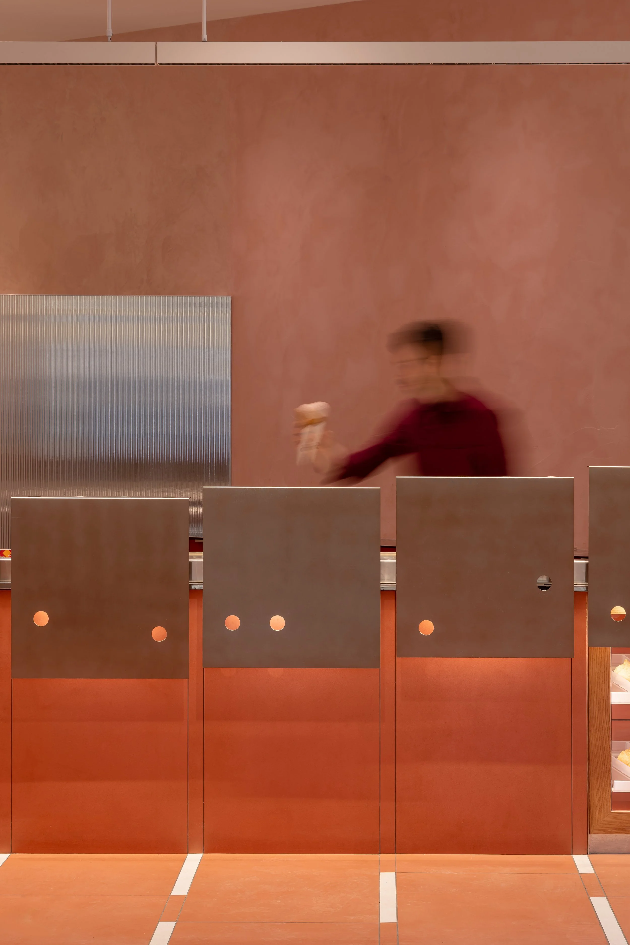 Contemporary ice cream shop interior with warm terracotta walls, fluted mirrored partial height walls, stainless steel counter panels, and blurred staff serving ice cream.