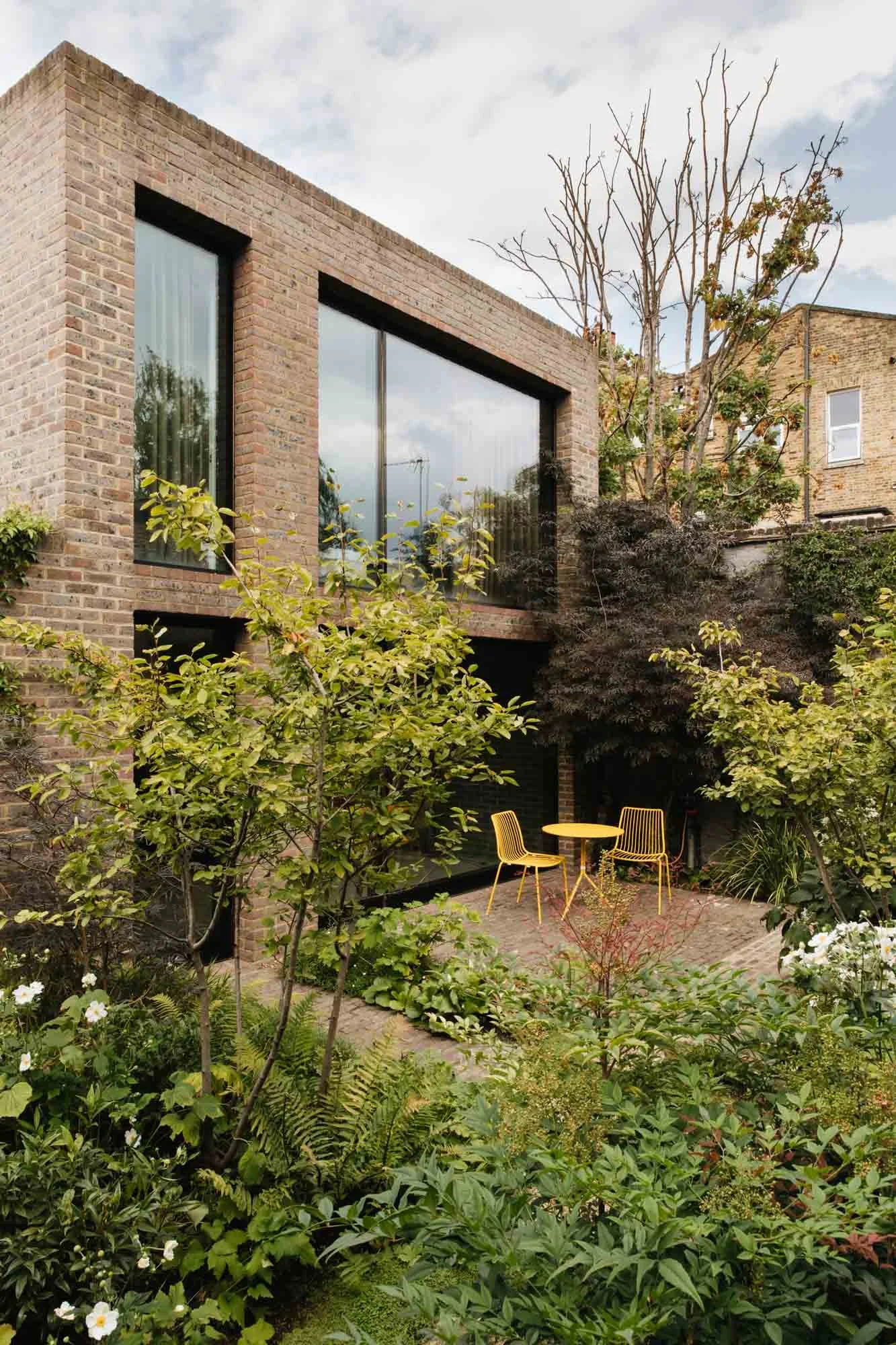A modern brick house with large windows and a small outdoor yellow table with three yellow chairs, surrounded by lush greenery and trees in a garden setting. Non Morris garden design