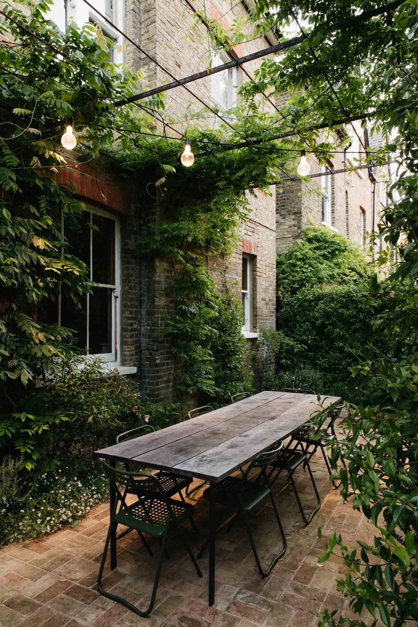 An outdoor brick patio with a wooden table and black chairs, surrounded by green foliage and vines, illuminated by string lights Sarah Price designed garden