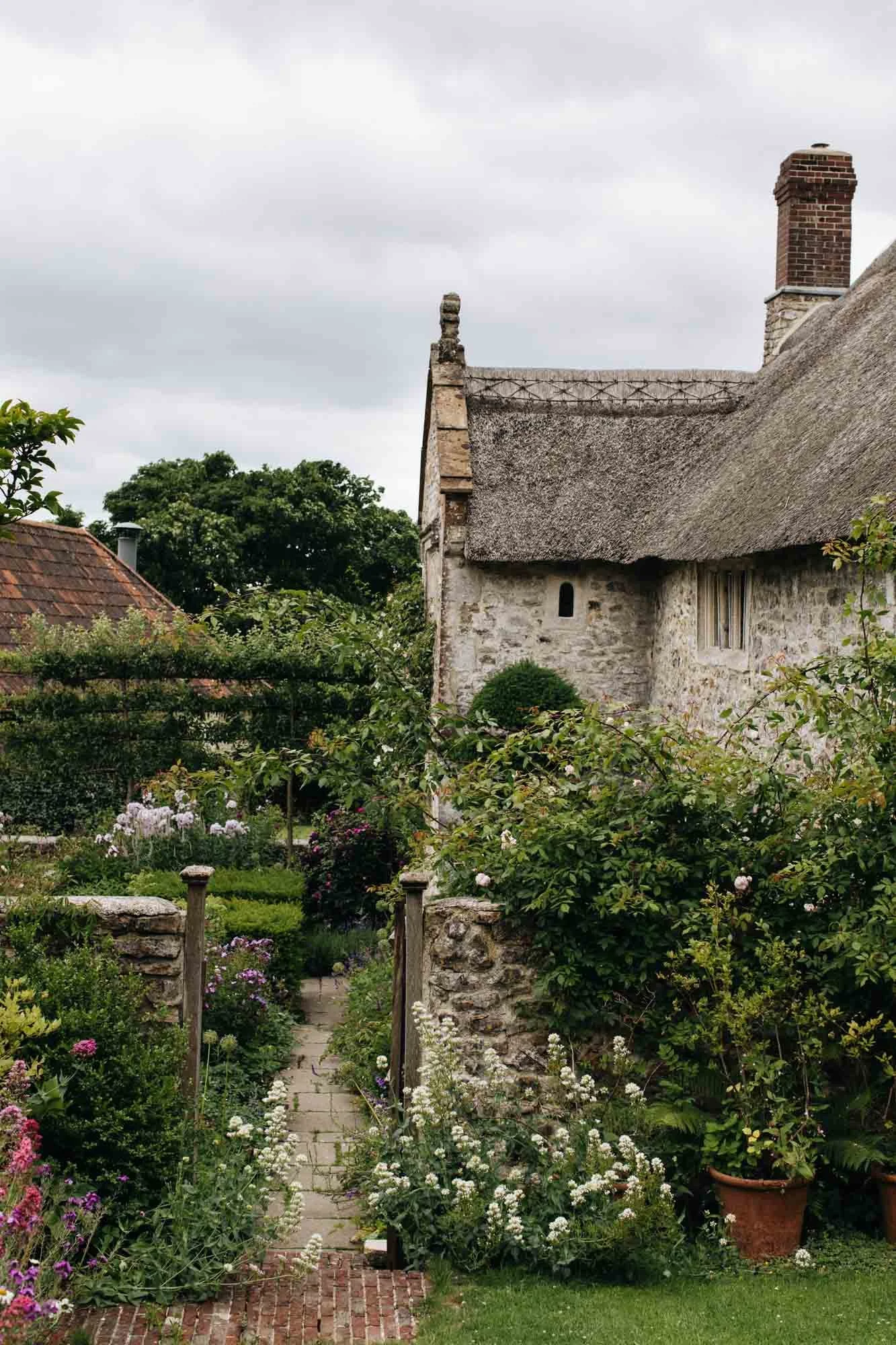 A quaint stone cottage with a thatched roof, chimney, and small window, surrounded by a lush garden with colorful flowers and a stone pathway. South Wood Farm by Arne Maynard