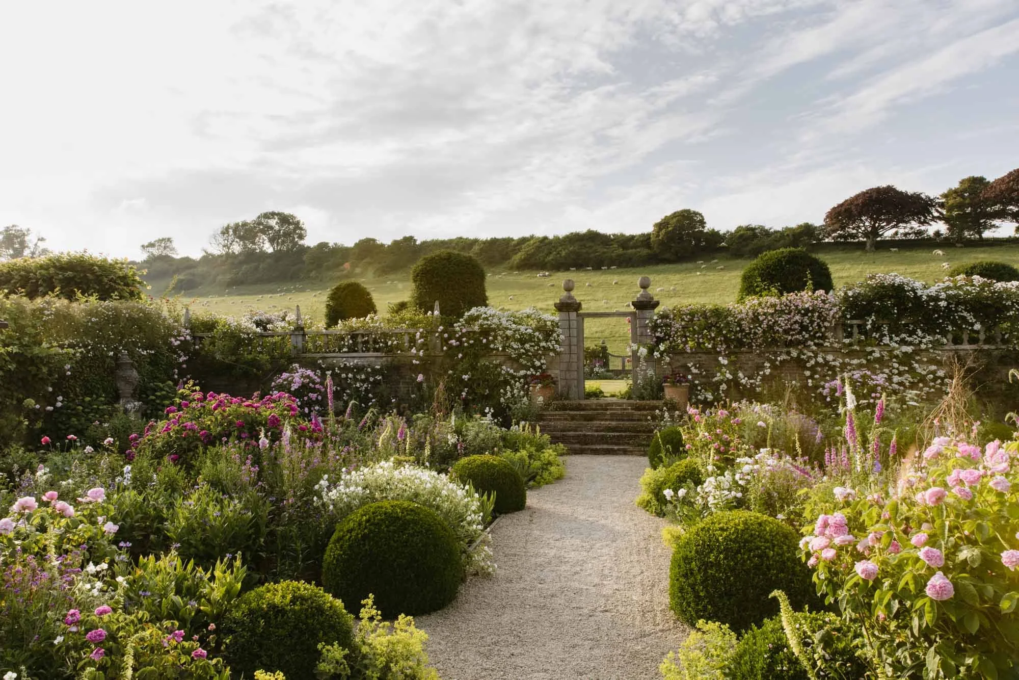 A lush garden with blooming flowers along a gravel pathway leading to a stone gate, with a green rolling hill and trees in the background under a partly cloudy sky. Dorset Walled Garden in Somerset