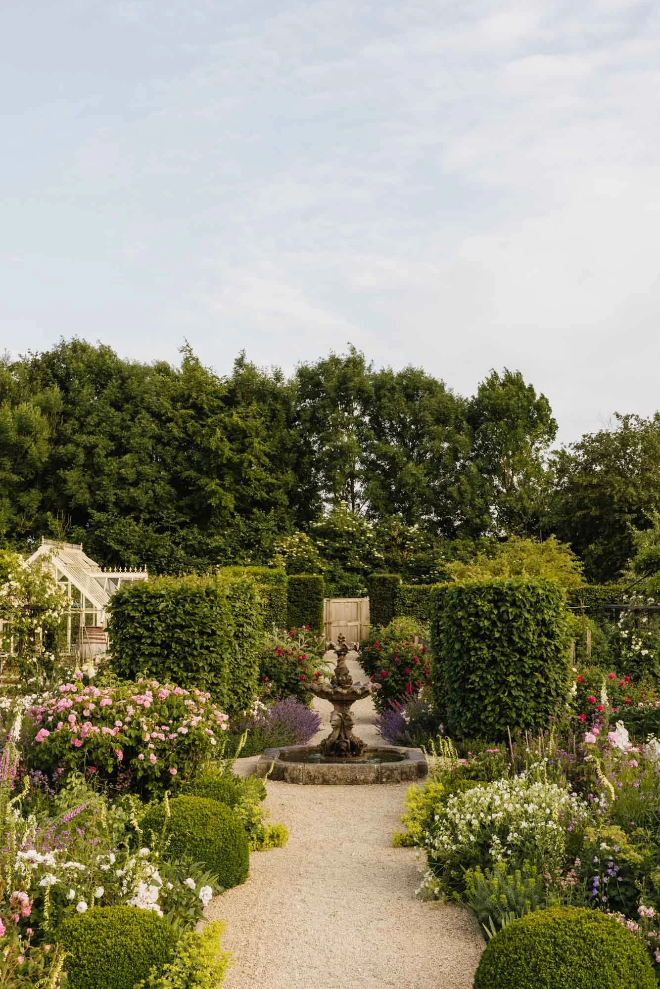 A garden with a gravel pathway, lush green bushes and flowering plants on either side, a decorative fountain at the center, surrounded by trees and shrubbery, under a cloudy sky. Dorset Walled Garden in Somerset
