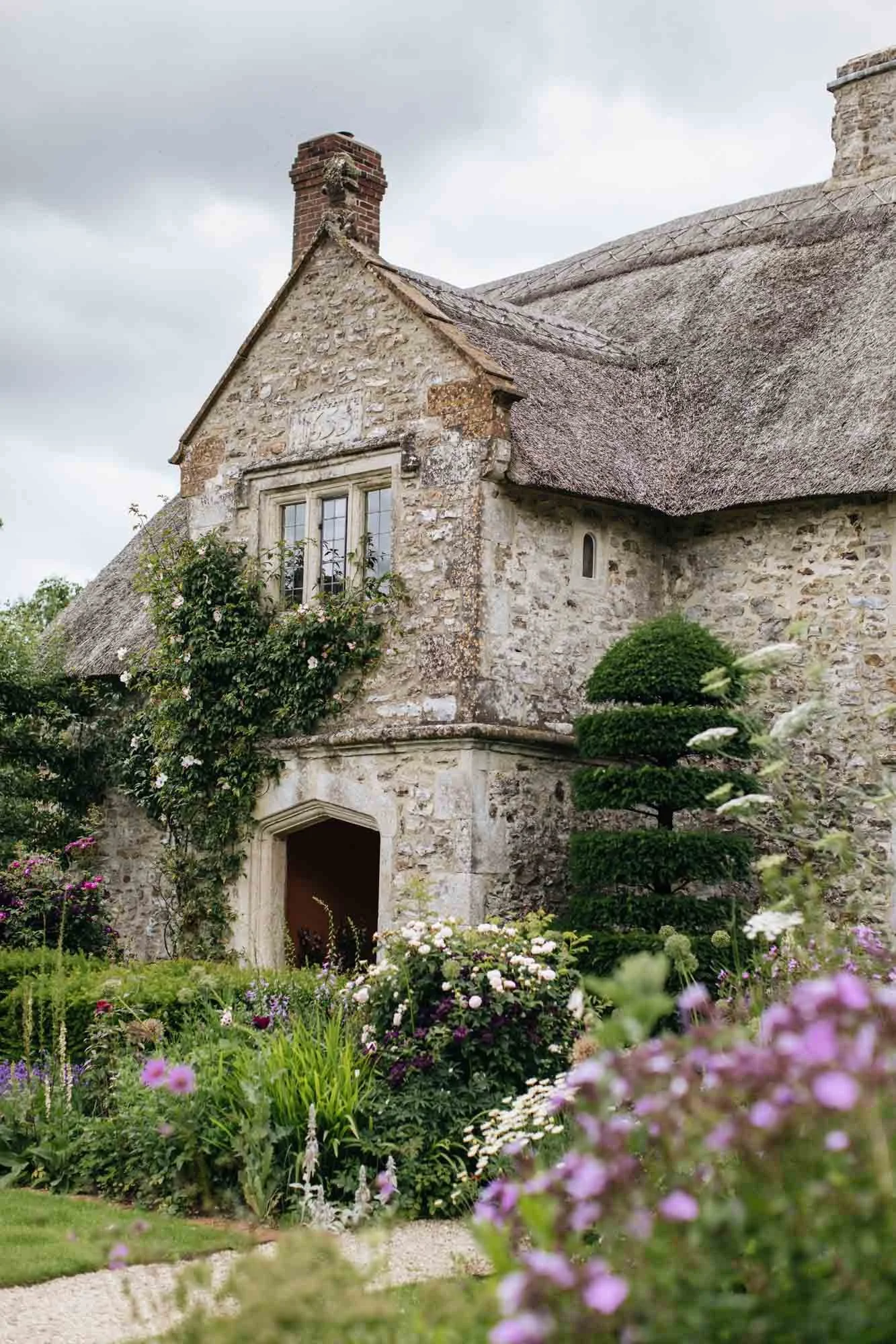 A stone cottage with a thatched roof, flowering garden, and lush greenery. South Wood Farm by Arne Maynard