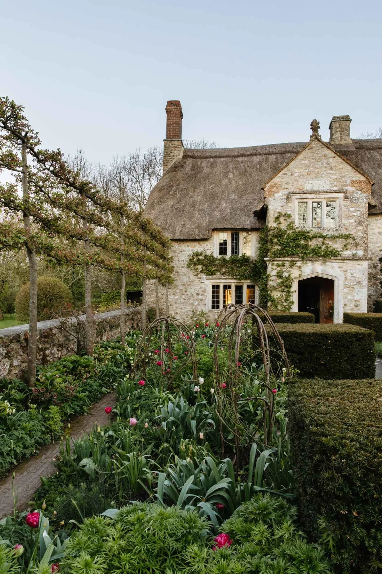South Wood Farm with a thatched roof and climbing vines, surrounded by lush garden with blooming flowers and neatly trimmed hedges.