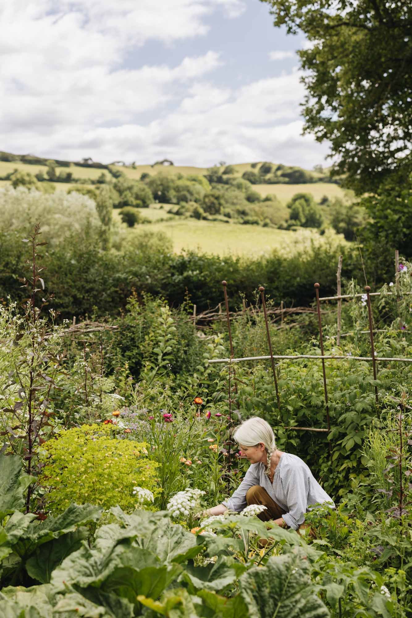 Alison Jenkins at Damson Farm tending to a lush garden with various plants and flowers, surrounded by green landscape and rolling hills under a partly cloudy sky.